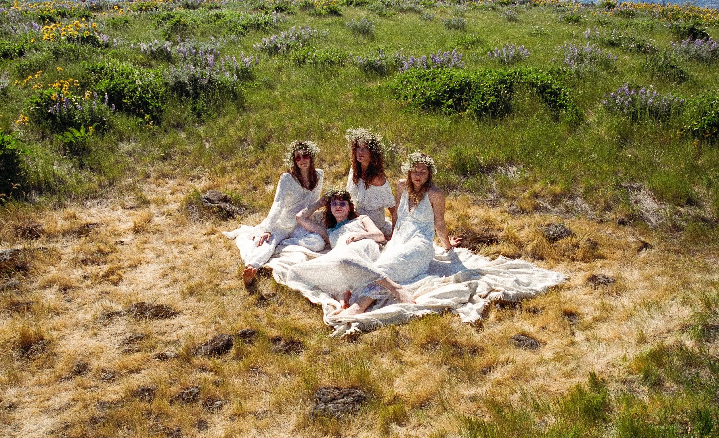Four women in white dresses with flower crowns sitting on a blanket in a grassy field with yellow and purple flowers.