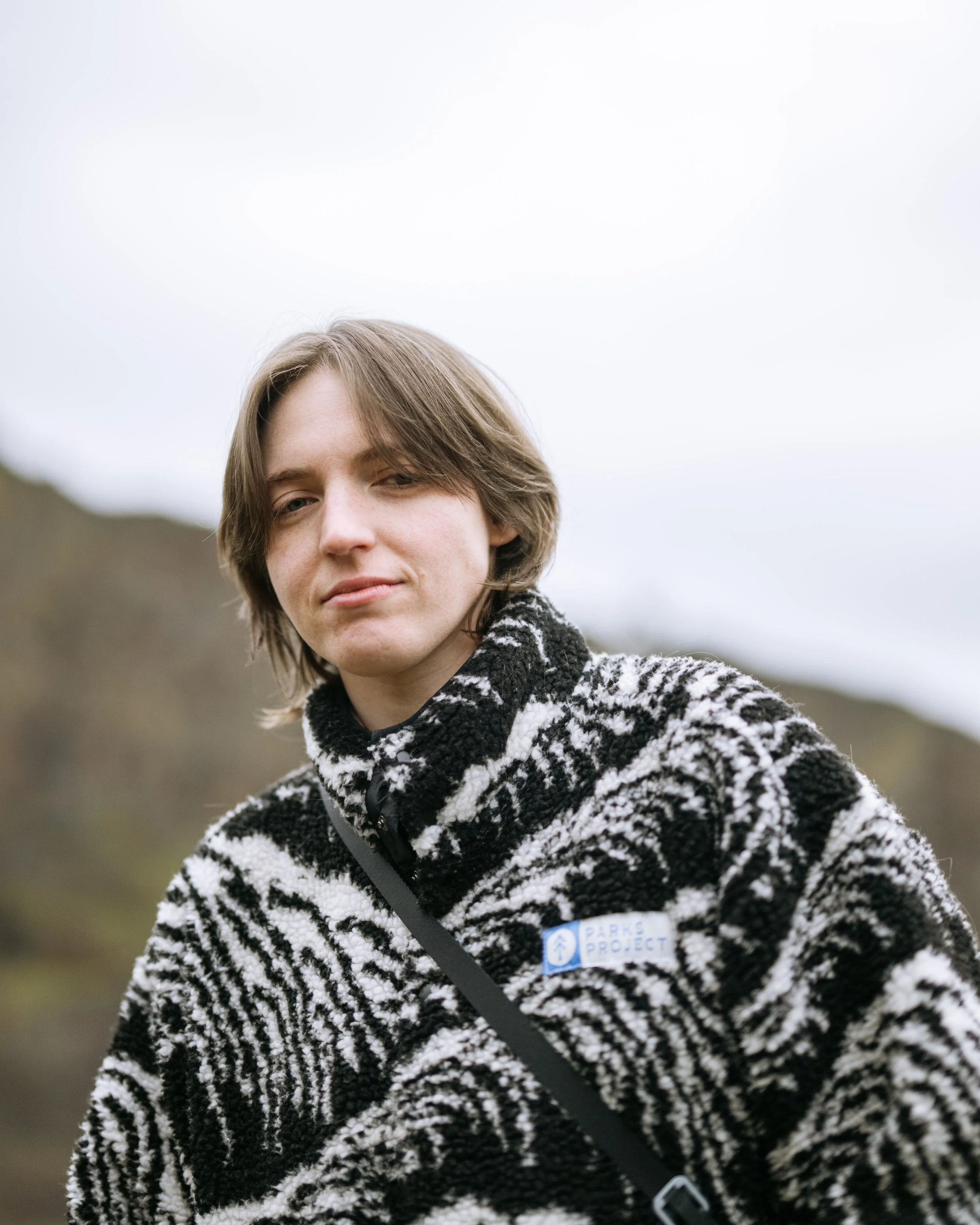 A young person with brown hair, wearing a black and white patterned sweater, smiling outdoors on a cloudy day.
