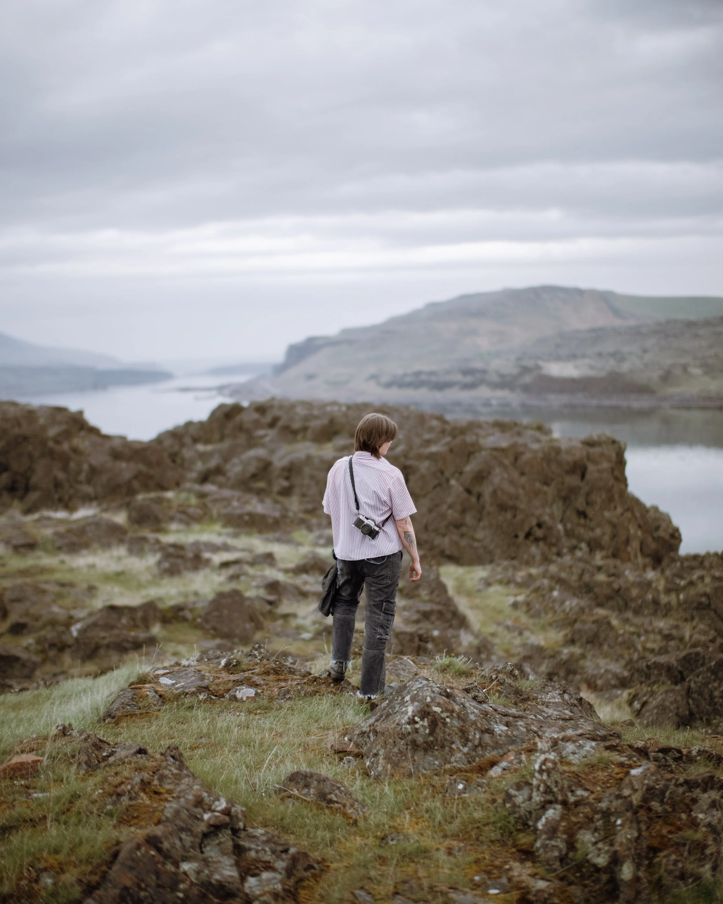 A person with short hair wearing a striped shirt and carrying a camera walks through rocky terrain with green grass, overlooking a foggy coastline with cliffs in the distance under a cloudy sky.