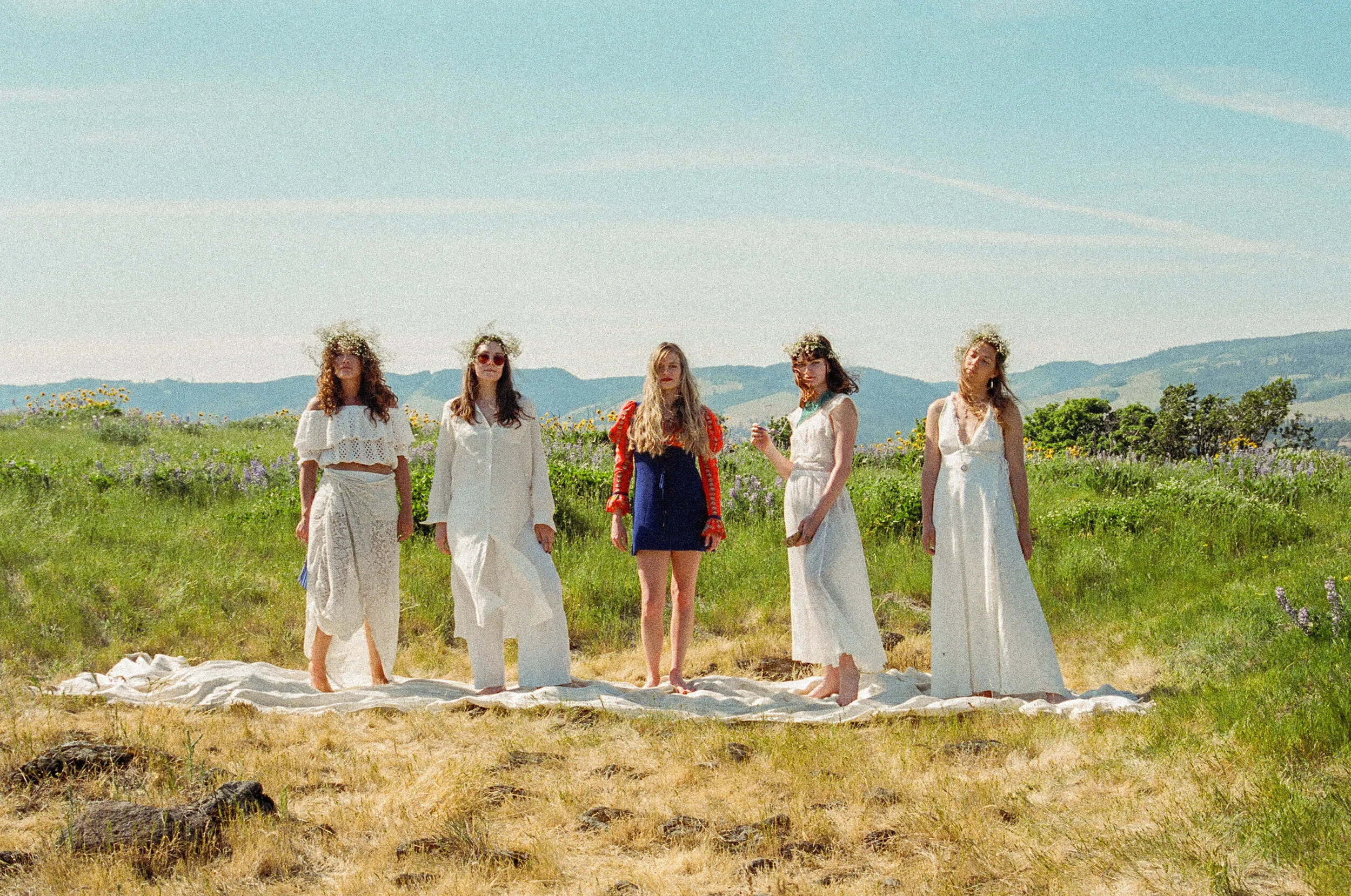 Group of five women standing in a field during daytime, some wearing flower crowns and white dresses, with rolling hills and blue sky in the background.