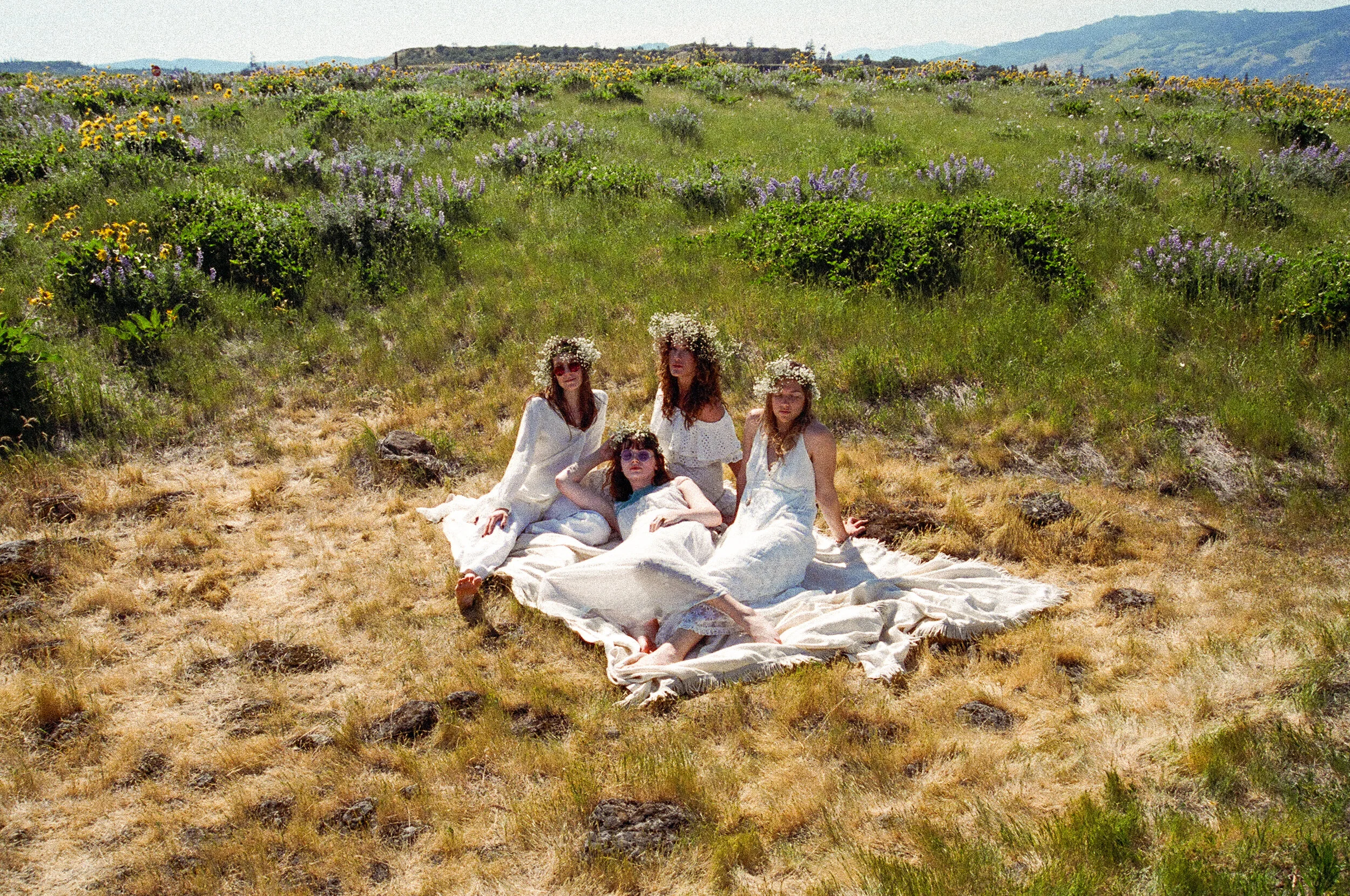 Four women with flower crowns and white dresses sitting on a blanket in a grassy field with purple and yellow flowers, hills in the background, during daytime.