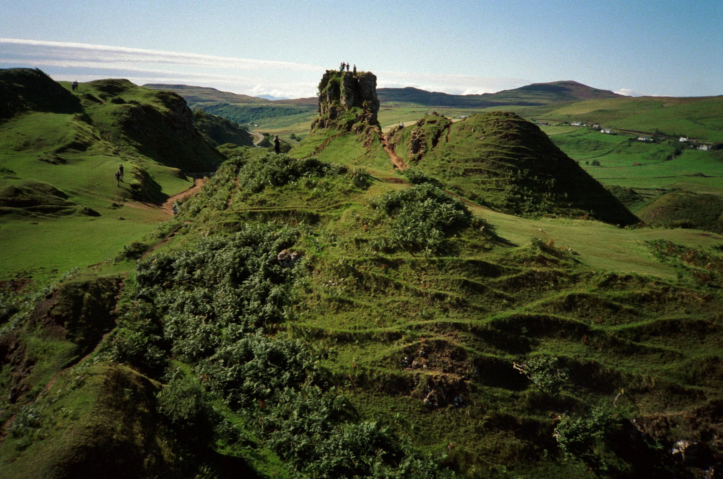 Green rolling hills with a tall rocky outcrop at the center, topped with a group of people, and a few more people walking along a dirt path on the hillside. Houses are visible in the distance under a blue sky with clouds.