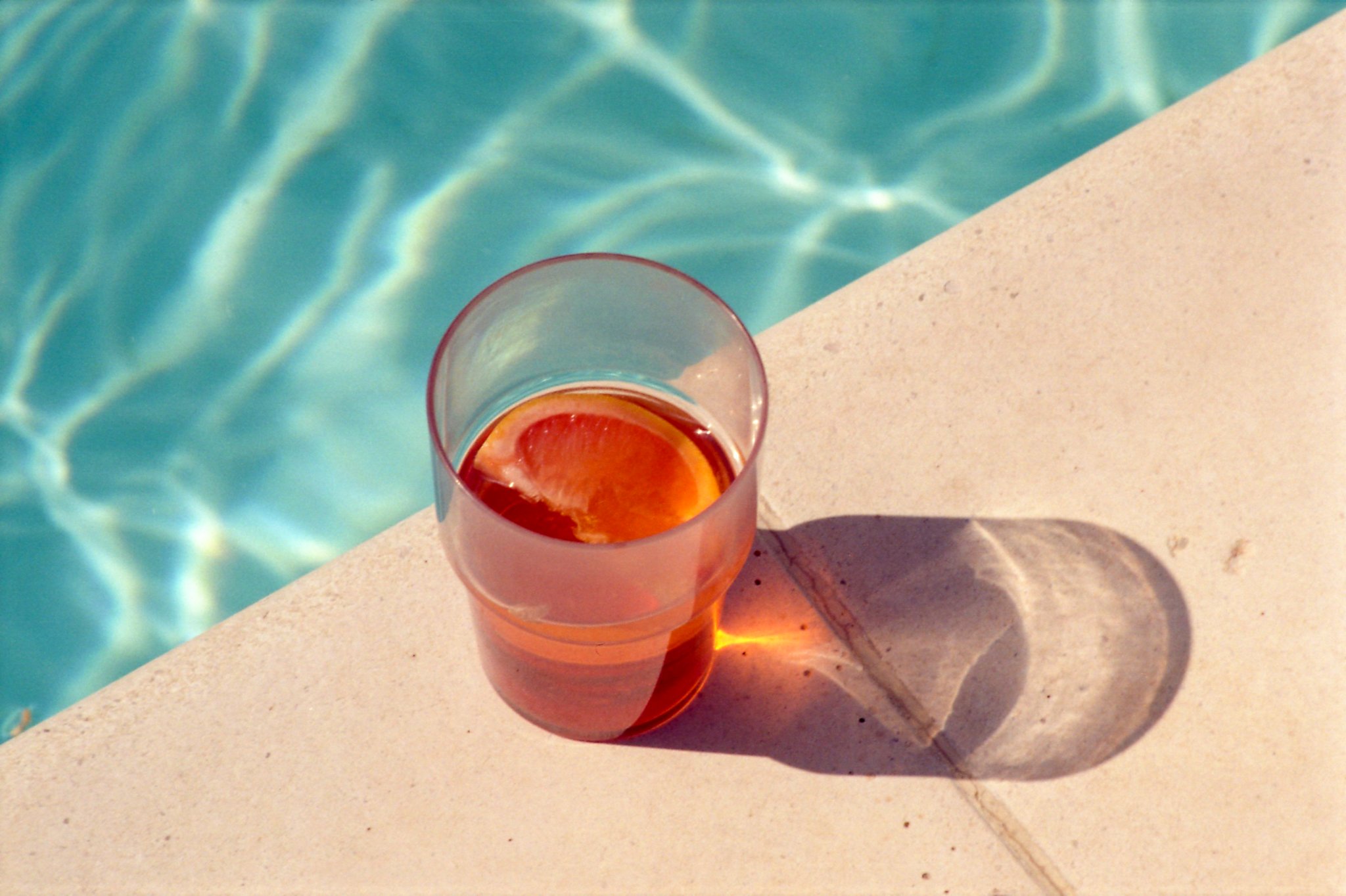 A tall glass of pink iced tea with ice and a slice of orange, sitting on a poolside concrete surface near a swimming pool with clear blue water.