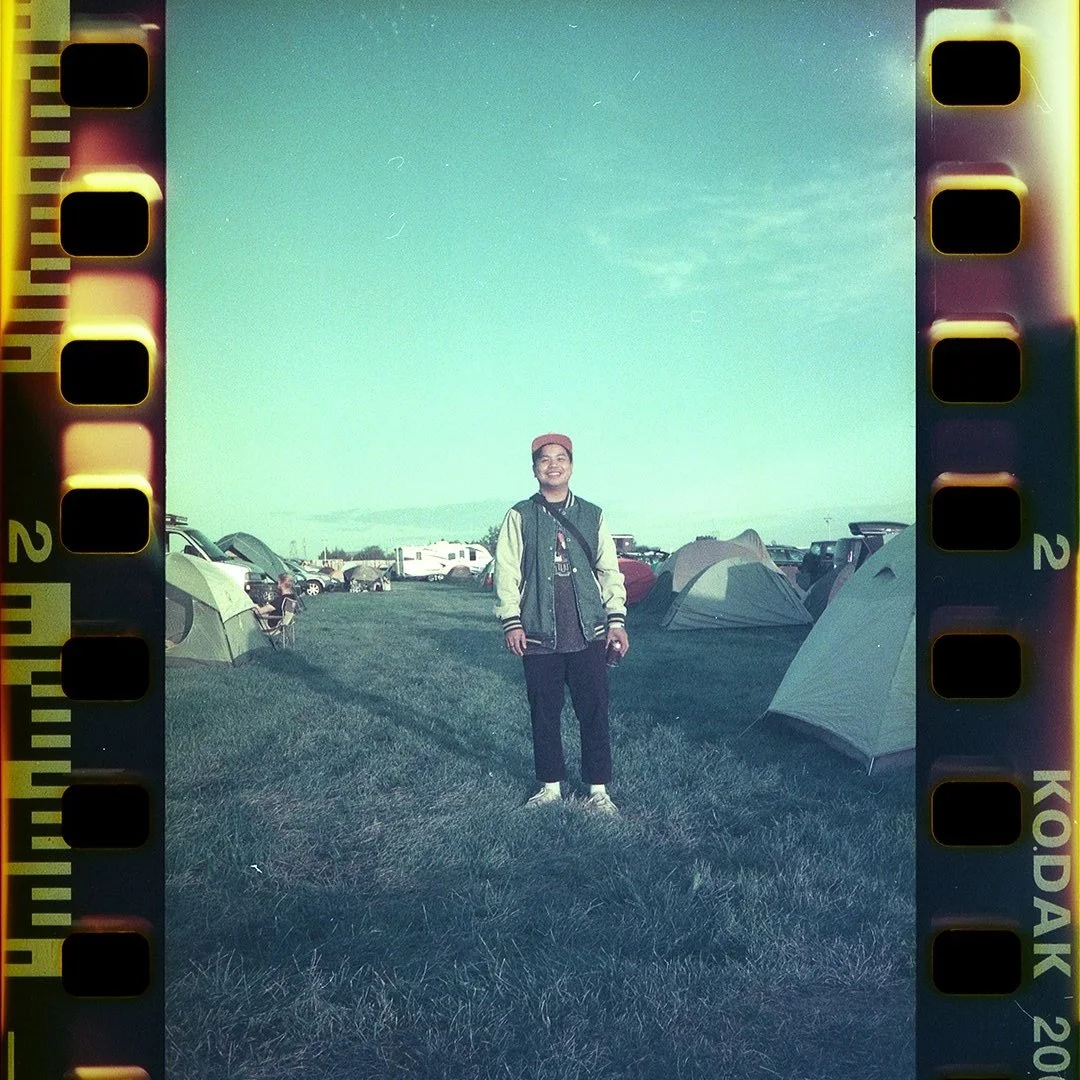 A young man standing on grass at a camping site with tents and cars in the background, under a light green sky.