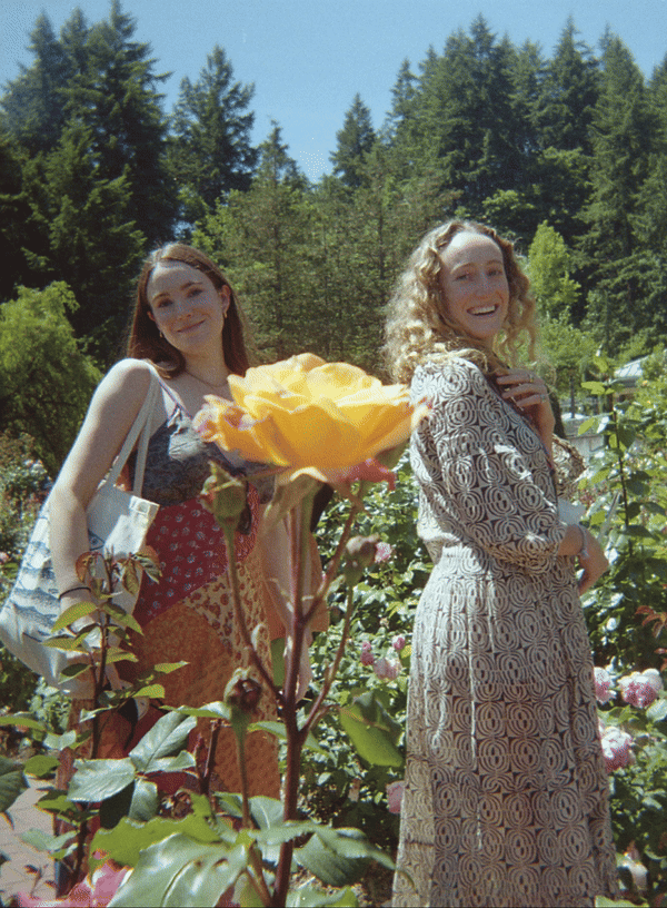 Two women standing in a rose garden with children in the background, smiling and posing for the camera on a sunny day.