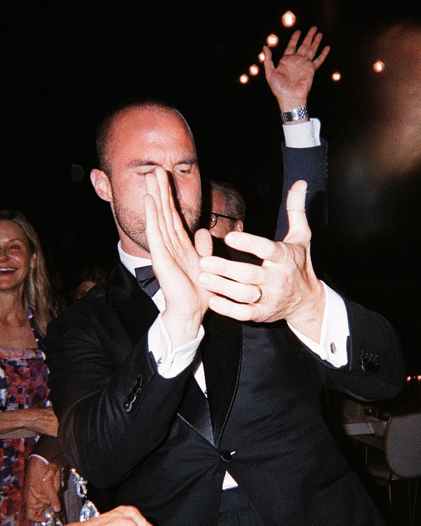 A man in a tuxedo dancing at an event with other people in the background, some smiling.