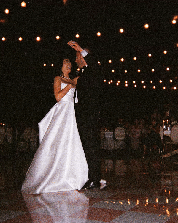 A bride and groom having their first dance at a wedding reception under hanging string lights.