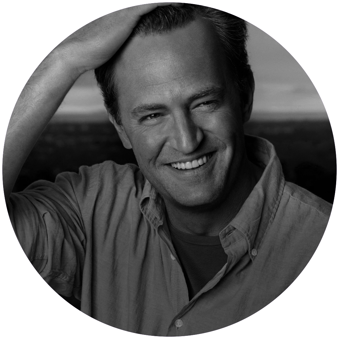 Black and white close-up of a smiling man with short hair, wearing a casual shirt, outdoors with a blurred landscape background.