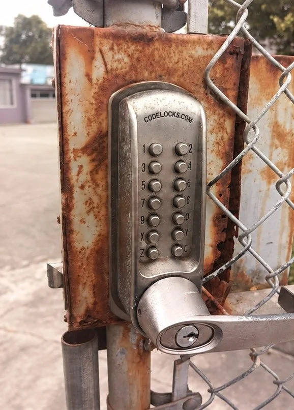 Rusted steel lock box on a chain-link gate before repair, with a keypad lock
