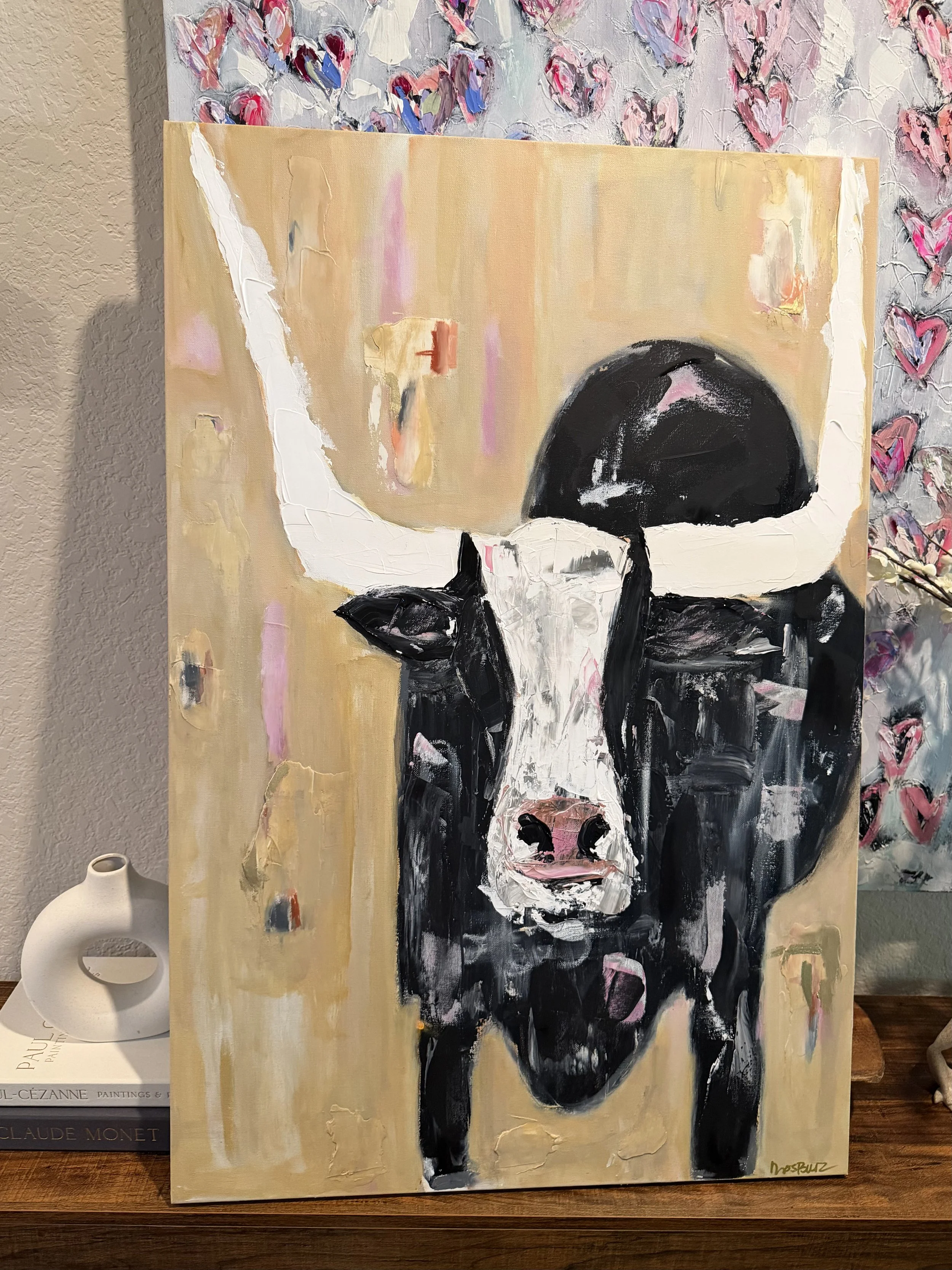 A portrait painting of a cow with black and white fur, set against a beige background with pink and purple abstract accents, signed in the bottom right corner.