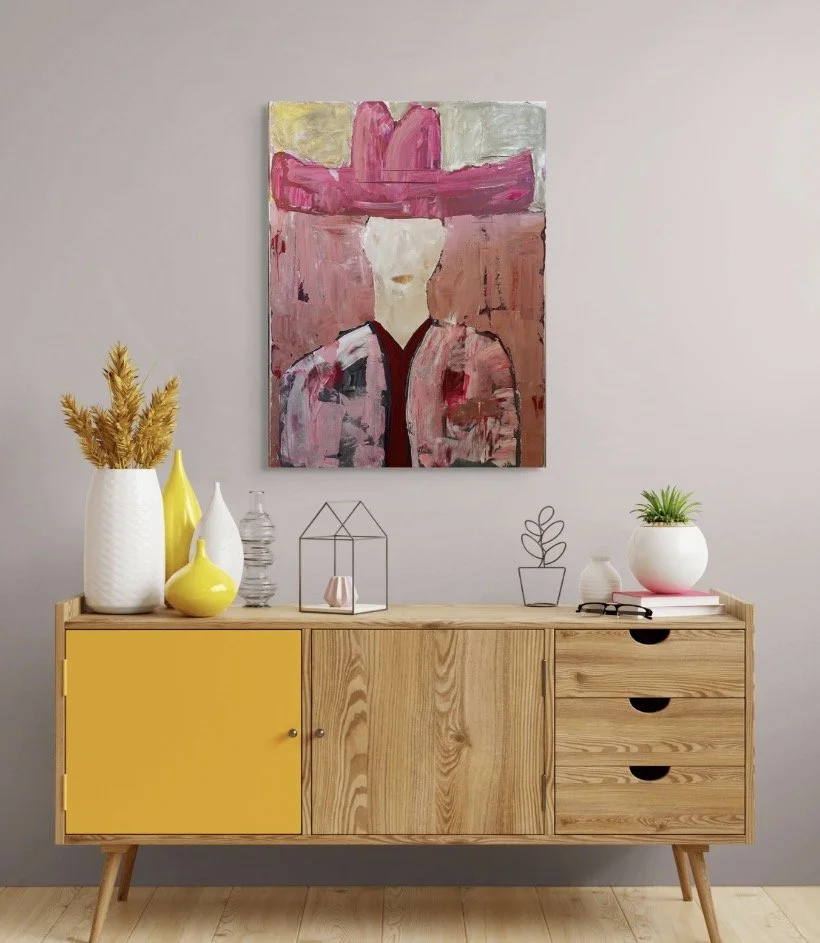Decorative wooden sideboard with yellow color accents, topped with vases and plants, hanging abstract painting of a figure with a pink hat on a gray wall.