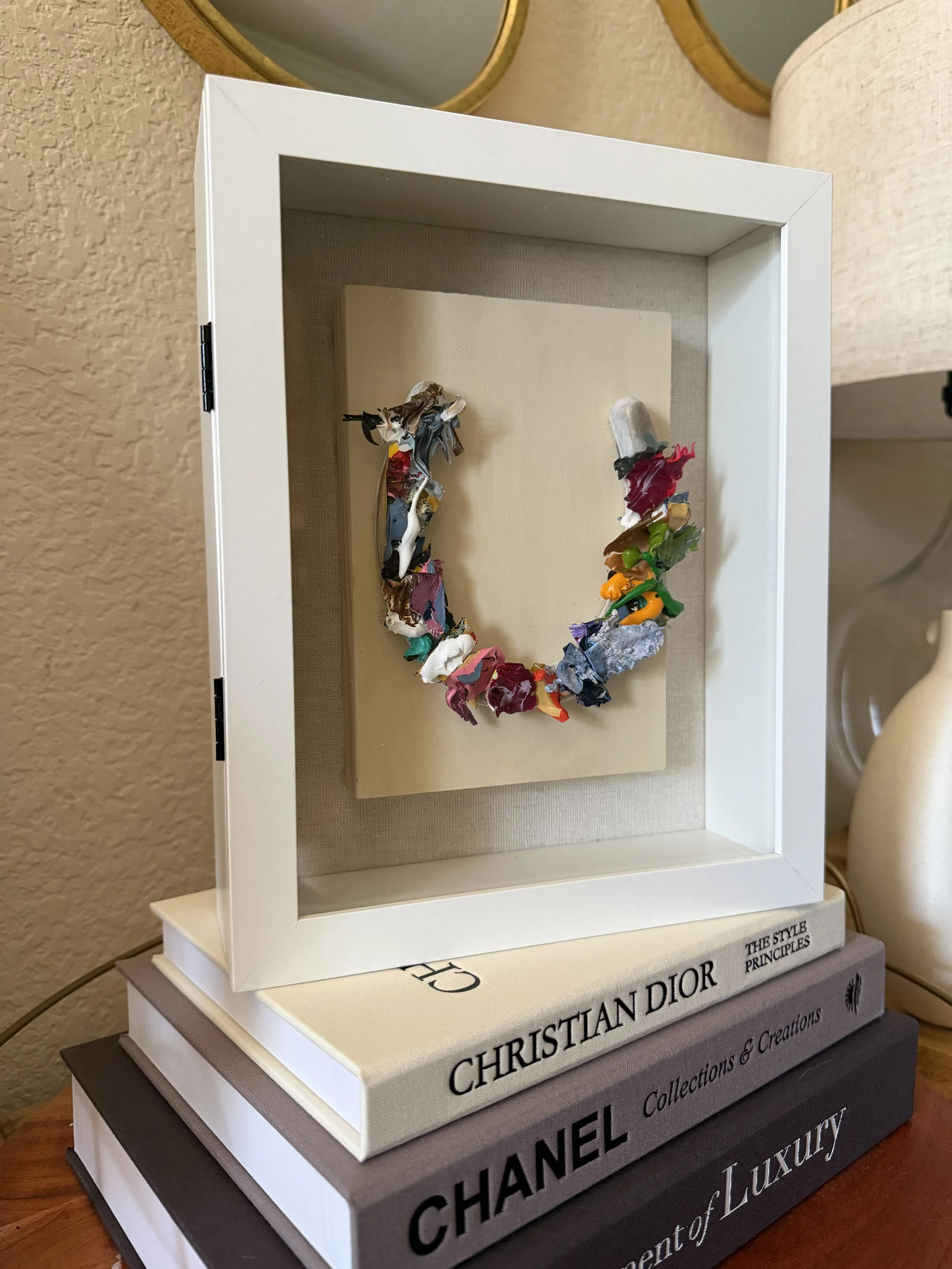 A framed art piece with colorful, textured abstract material arranged in a circular pattern, placed on top of stacked books about fashion brands and luxury, with a beige wall and decorative mirrors in the background.