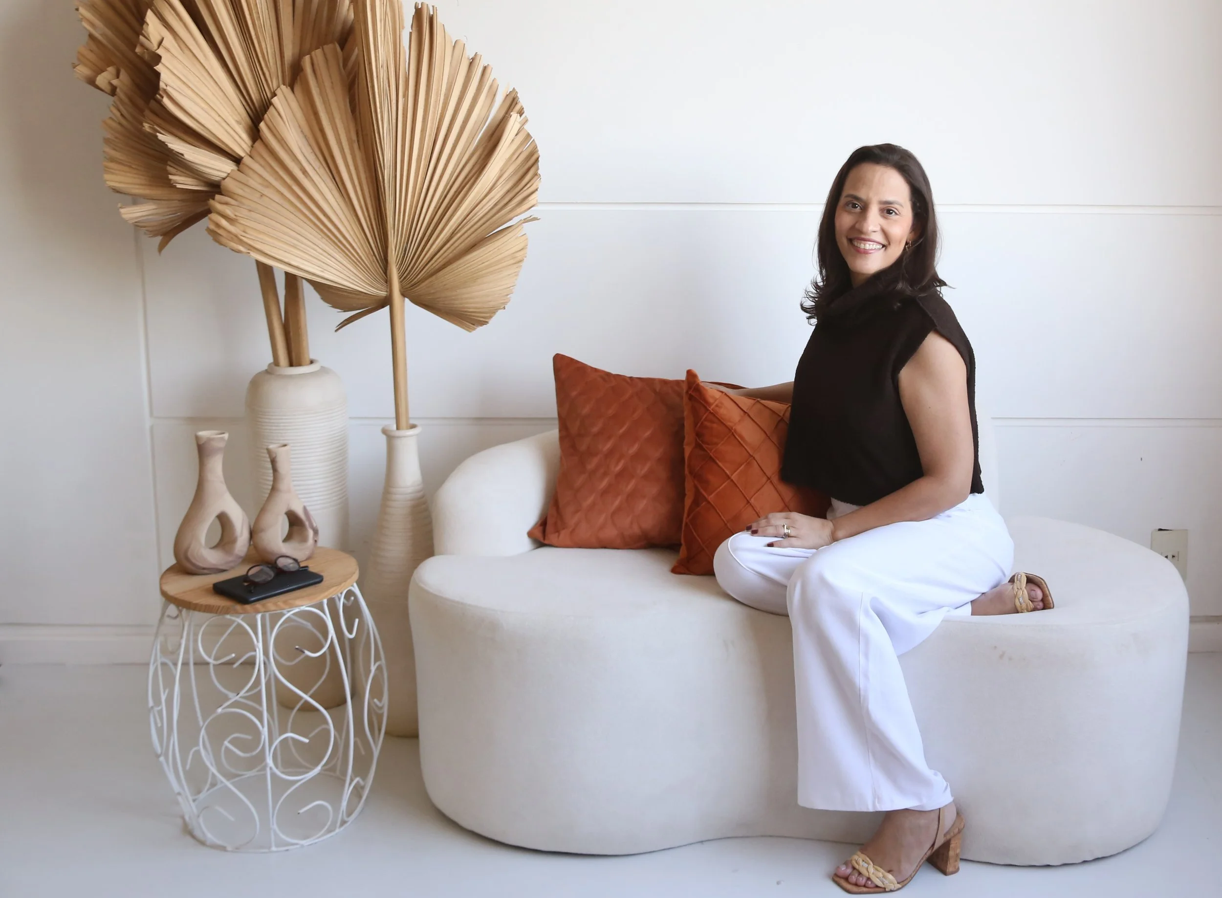 Mulher sorridente, sentada em um sofá branco, segurando uma almofada laranja. Ao seu lado, uma mesa de ferro branca com óculos escuros, um celular e uma inscrição que diz 'love'. Atrás, decoração com vasos de cerâmica e uma grande obra decorativa de folhas secas.