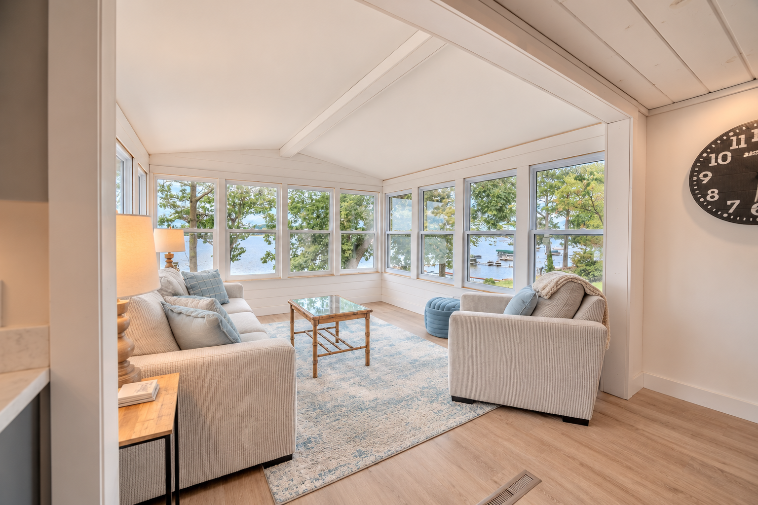 Bright lakeside sunroom with large windows, upholstered sofas and armchair, glass coffee table, blue ottoman, and area rug
