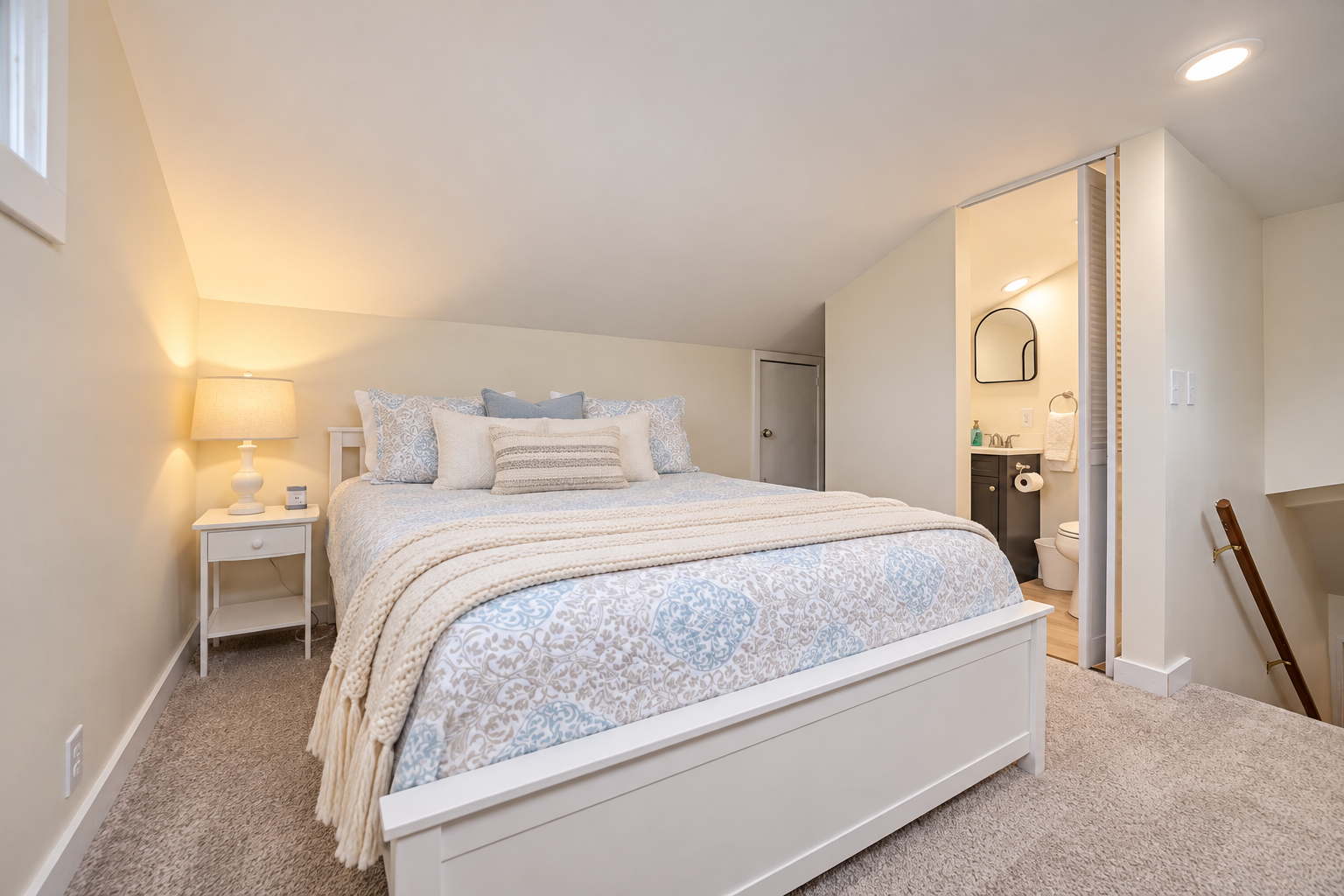 Bright, cozy bedroom with a white bed, blue-and-white patterned bedding, bedside table and lamp, and an adjoining bathroom.