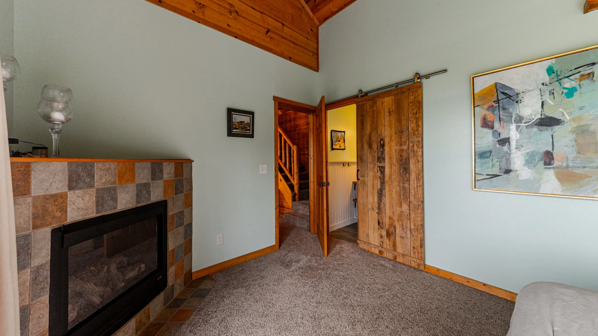 Cozy living room with tiled fireplace, exposed wood ceiling, barn door, staircase, and abstract wall art