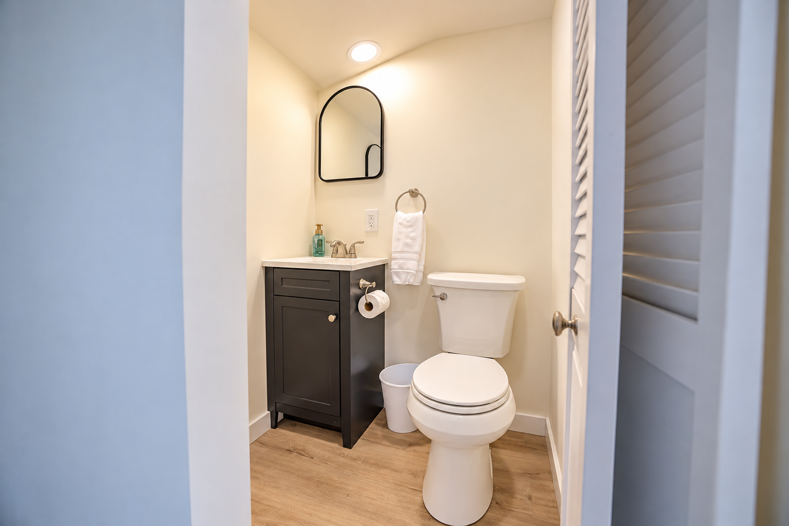 Small bathroom with a black vanity, white sink, wall mirror, toilet, towel ring, and wood-look flooring
