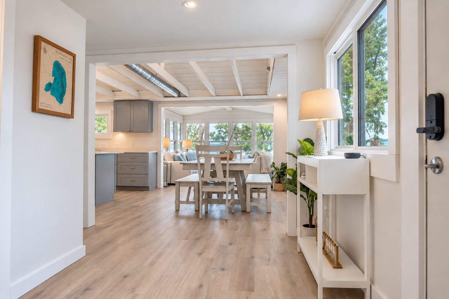 Bright modern open-plan home interior with dining table, gray kitchen cabinets, plants, large windows, and lake views