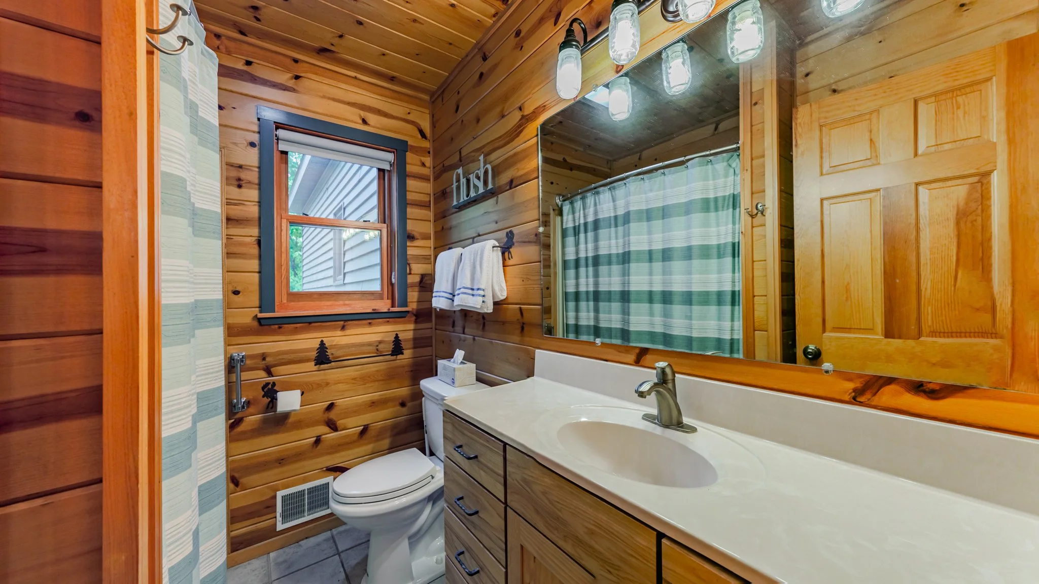 Rustic wood-paneled bathroom with a white vanity, large mirror, toilet, window, towels, and striped shower curtain