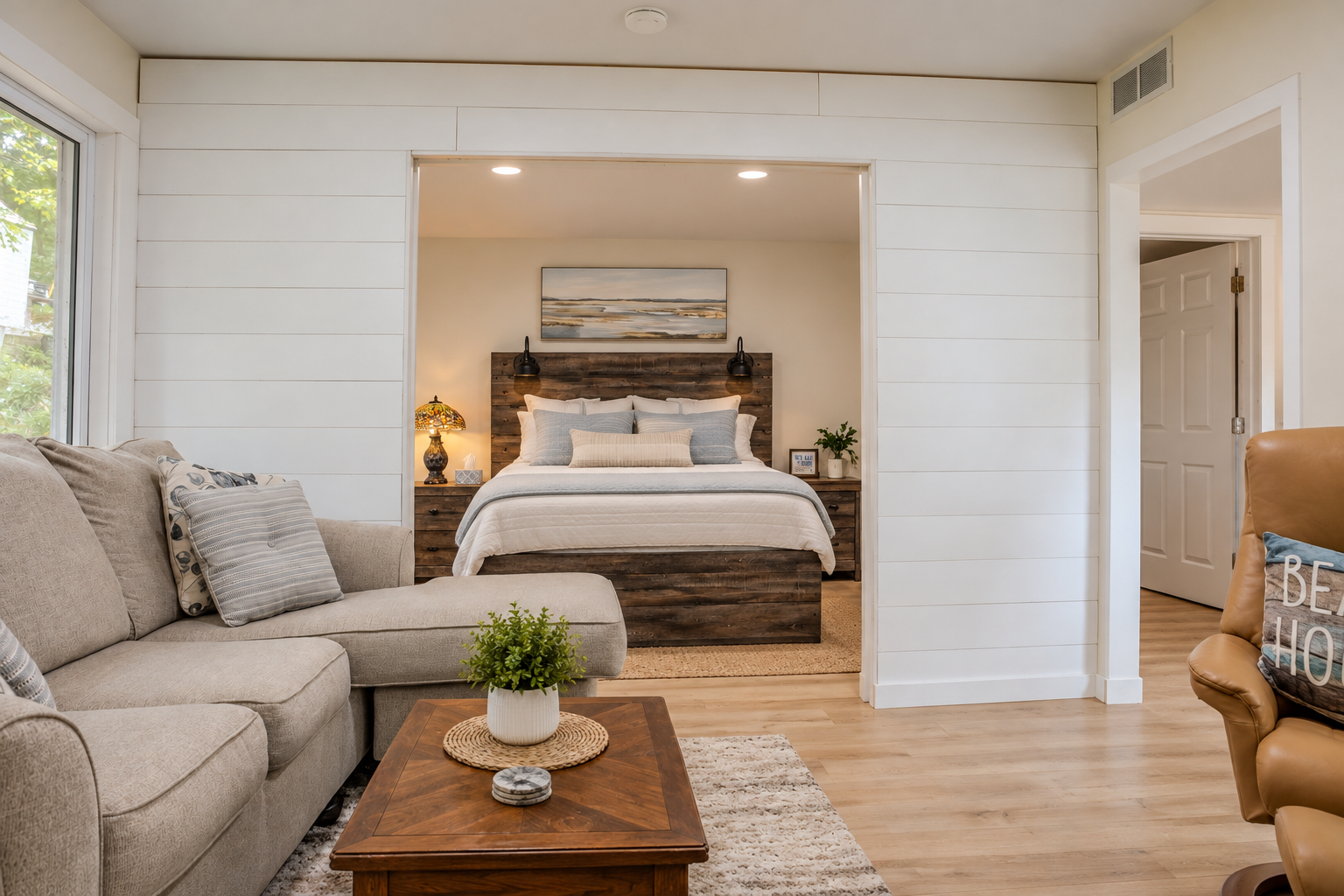 Cozy open-concept living room and bedroom with a white sectional sofa, wood coffee table, upholstered bed, and rustic wood headboard.