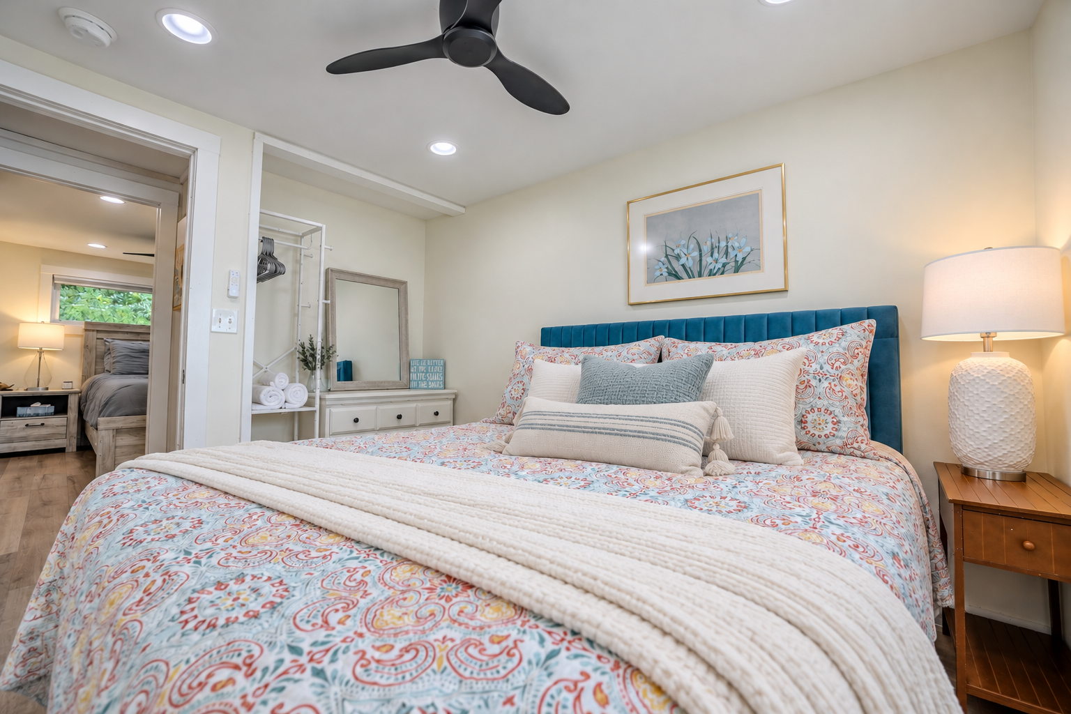 Bright coastal-style bedroom with a patterned comforter, teal upholstered bed, decorative pillows, bedside lamp, ceiling fan, and open closet