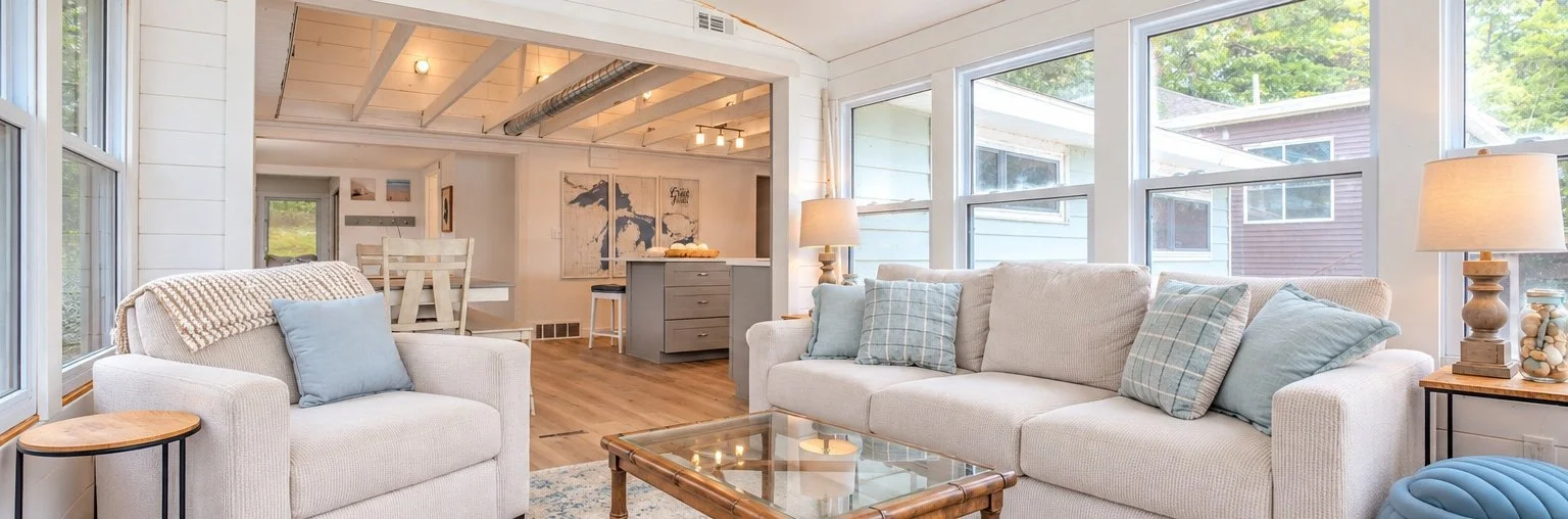 Bright coastal-style living room with white sofas, blue pillows, glass coffee table, large windows, and an open kitchen dining area.