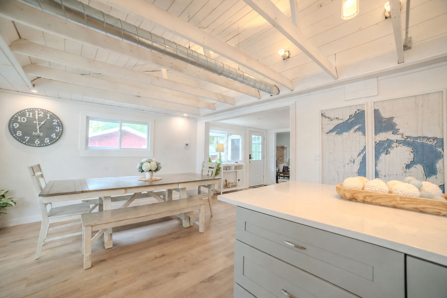 Bright white coastal-style kitchen and dining area with farmhouse table, benches, wall map artwork, large clock, and kitchen island.