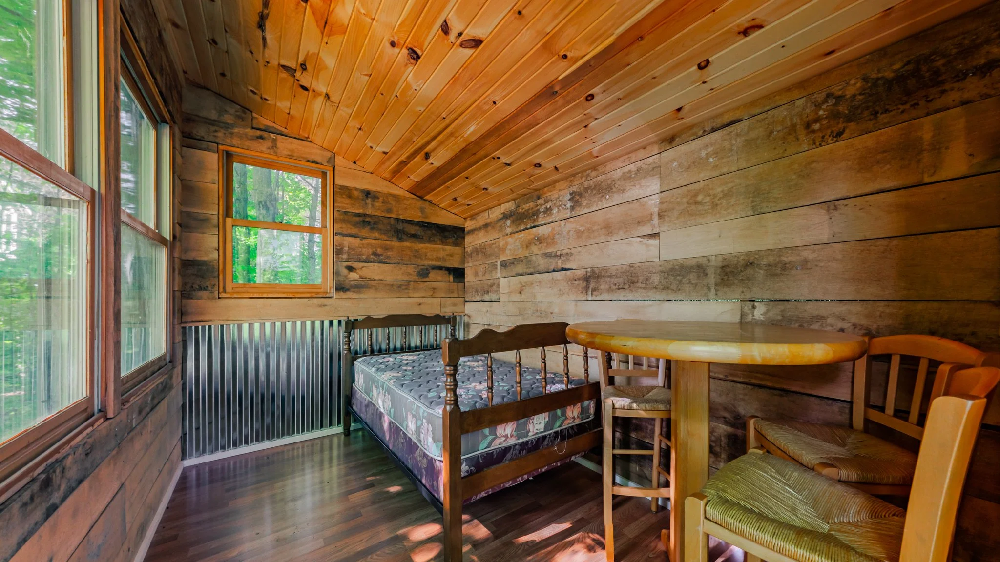 Rustic wood-paneled cabin room with a bed, round dining table, chairs, windows, and forest views