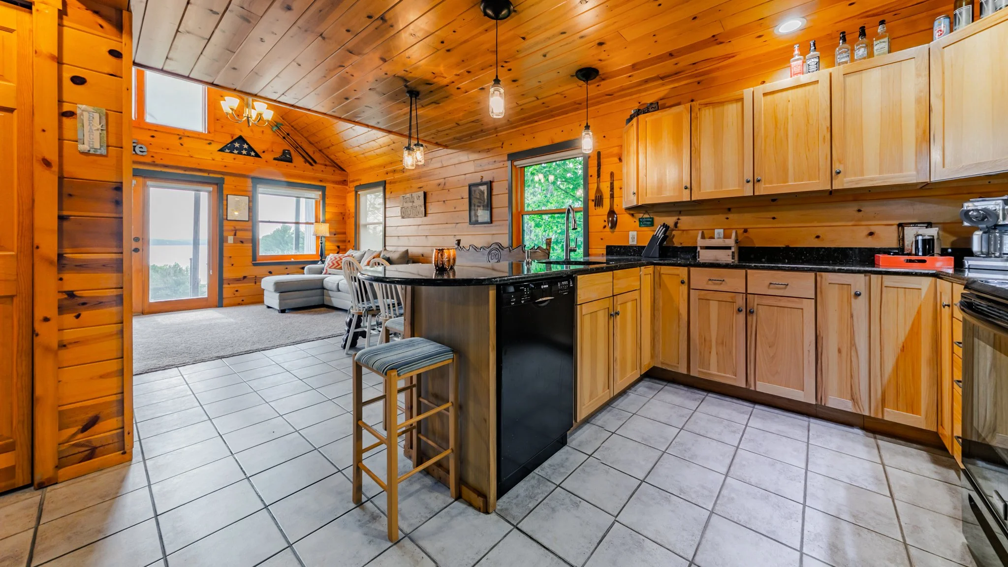 Rustic wood-paneled cabin kitchen with island, bar stools, wood cabinets, black countertops, and adjacent living room