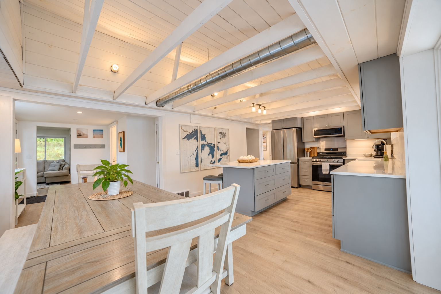 Bright farmhouse-style kitchen and dining area with exposed white beams, gray cabinets, stainless steel appliances, island, and wooden dining table.