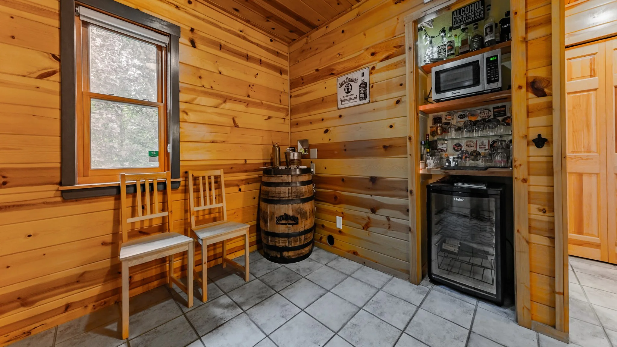 Rustic wood-paneled cabin bar area with two chairs, a whiskey barrel table, microwave, glassware, and beverage refrigerator.