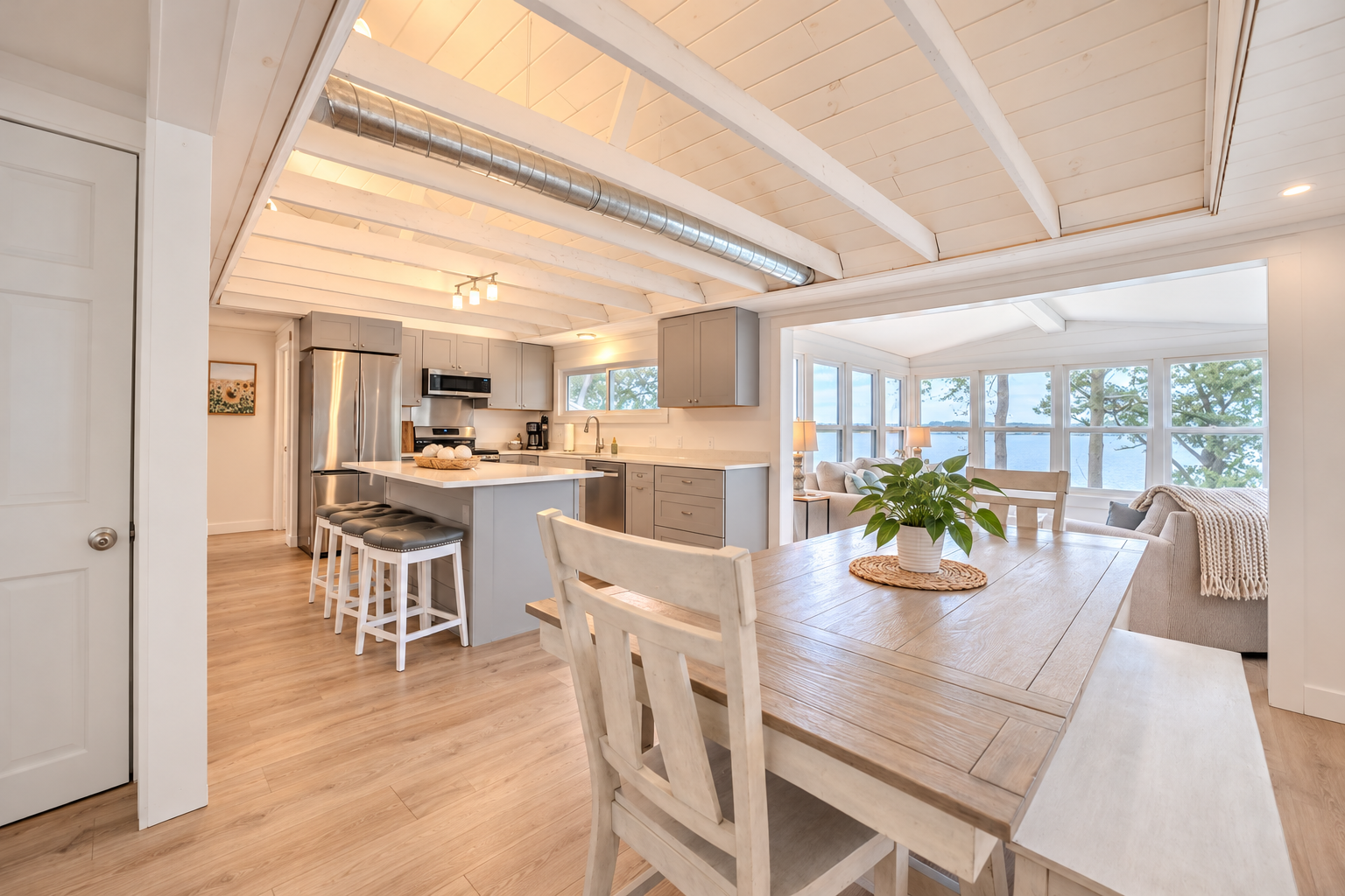 Bright open-concept kitchen and dining area with white beams, gray cabinets, island seating, and waterfront views through large windows