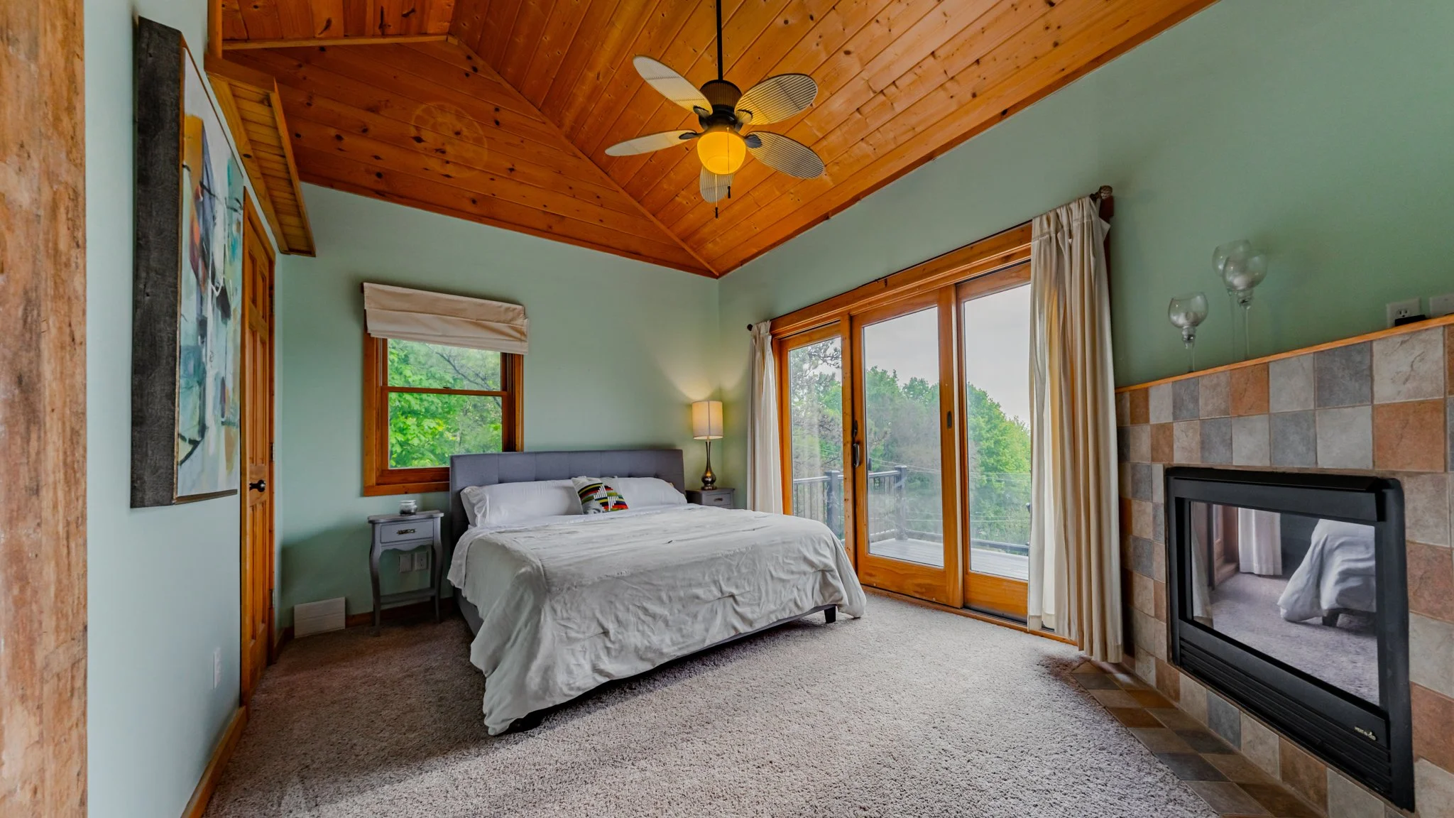 Cozy bedroom with a king-size bed, wood-paneled vaulted ceiling, balcony doors, and tiled fireplace.