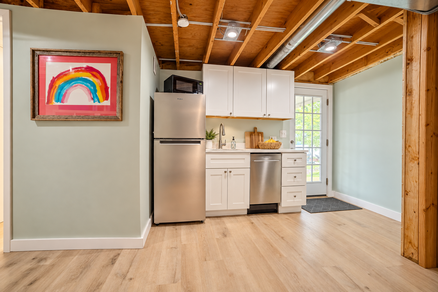 Bright compact kitchen with white cabinets, stainless steel appliances, exposed wood ceiling beams, and framed rainbow artwork