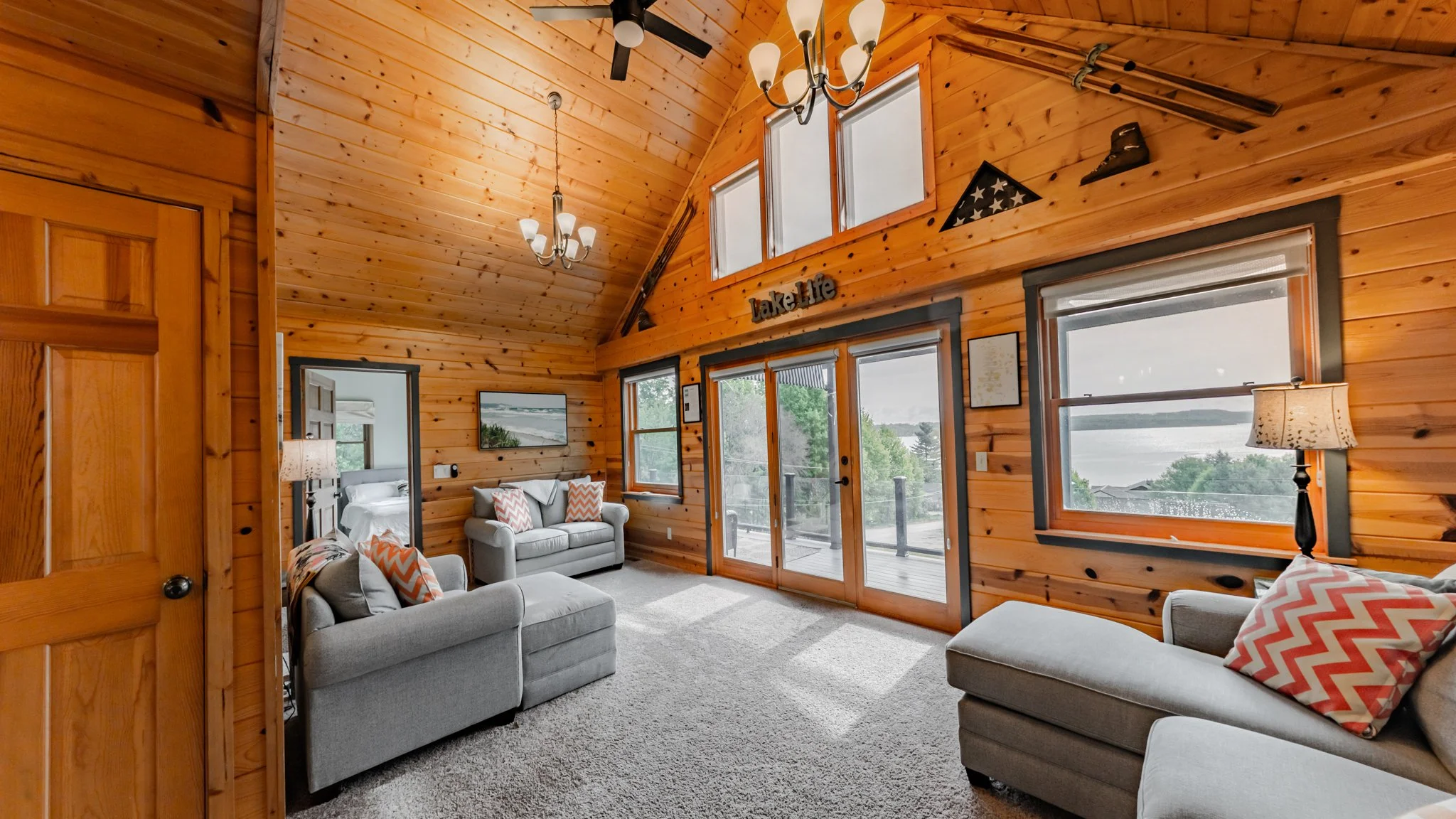 Rustic wood-paneled cabin living room with gray sofas, vaulted ceiling, large windows, and lake views