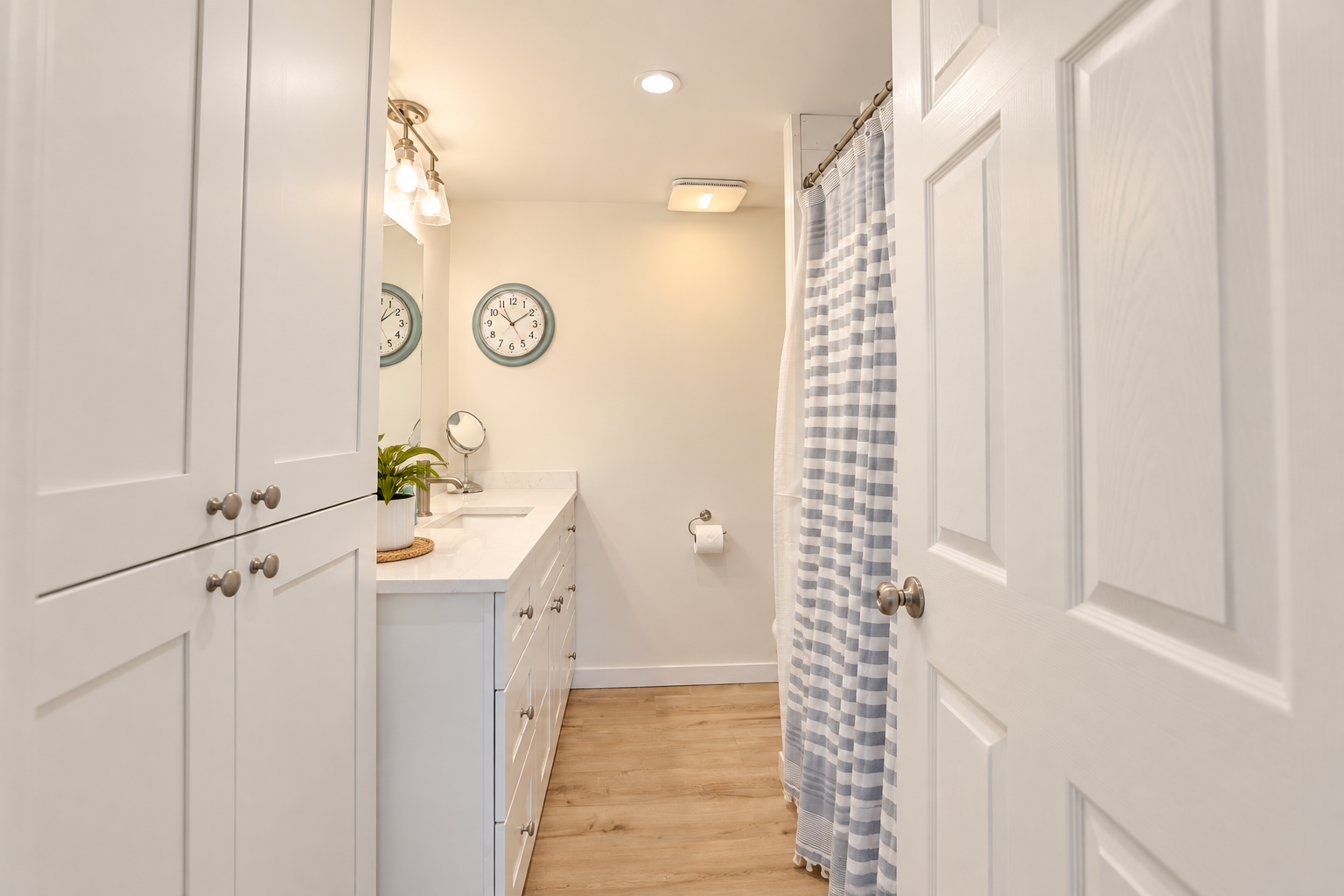 Bright white bathroom with vanity, mirrored cabinet, blue-and-white striped shower curtain, wall clock, and wood-look flooring