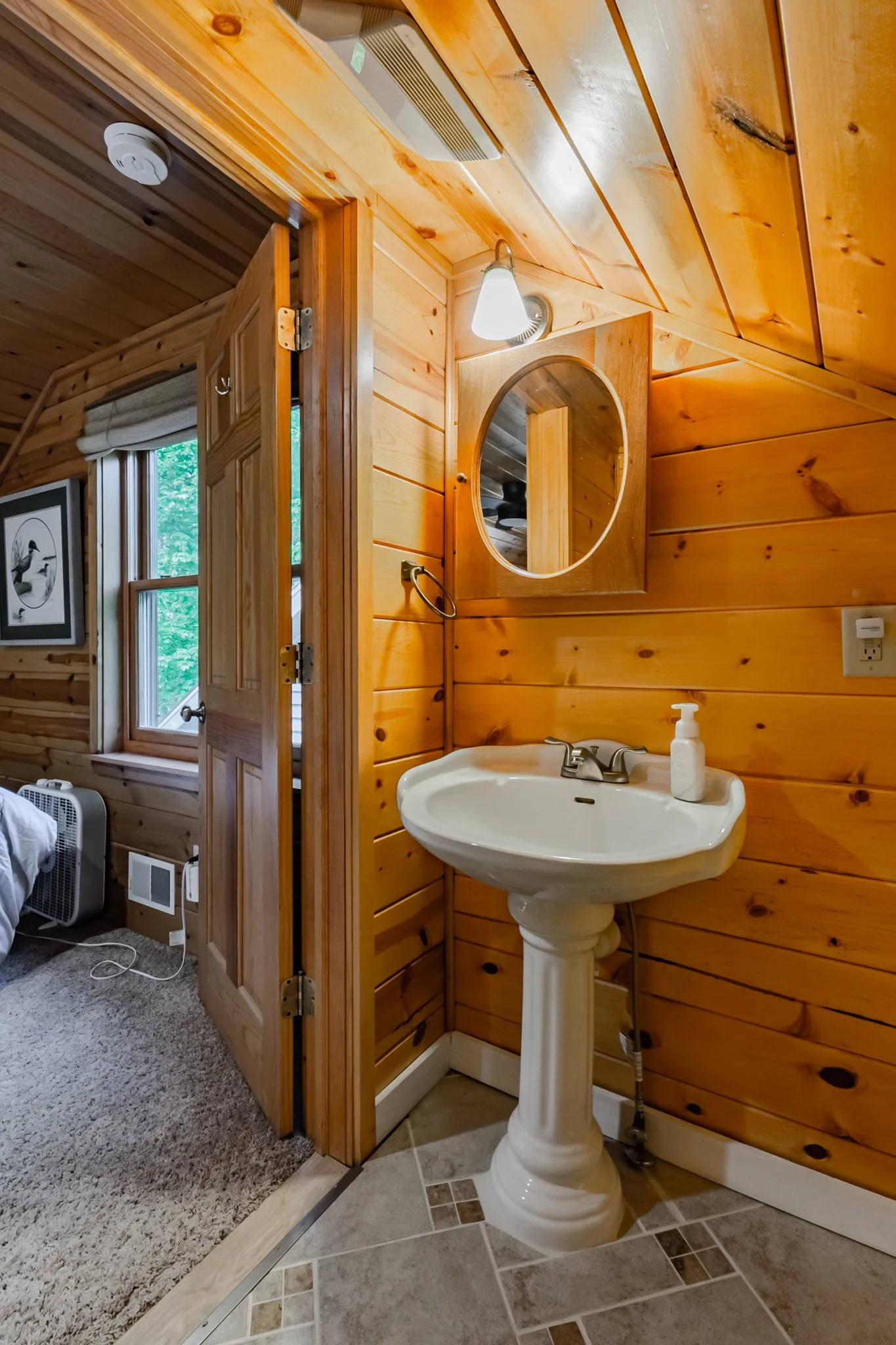 Rustic cabin bathroom with a white pedestal sink, oval mirror, wood-paneled walls, and adjoining bedroom