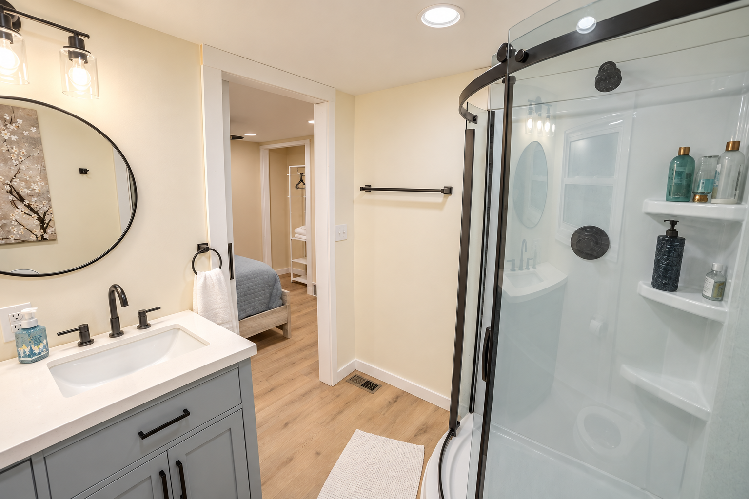 Bright modern bathroom with gray vanity, round mirror, glass shower enclosure, towel rack, and wood-look flooring