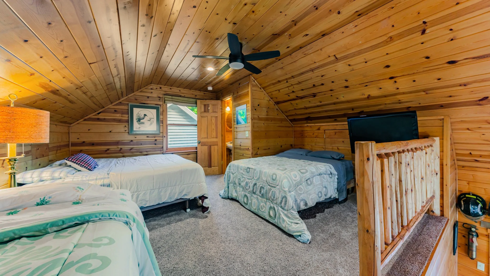 Rustic wood-paneled cabin bedroom with three beds, ceiling fan, television, and log railing