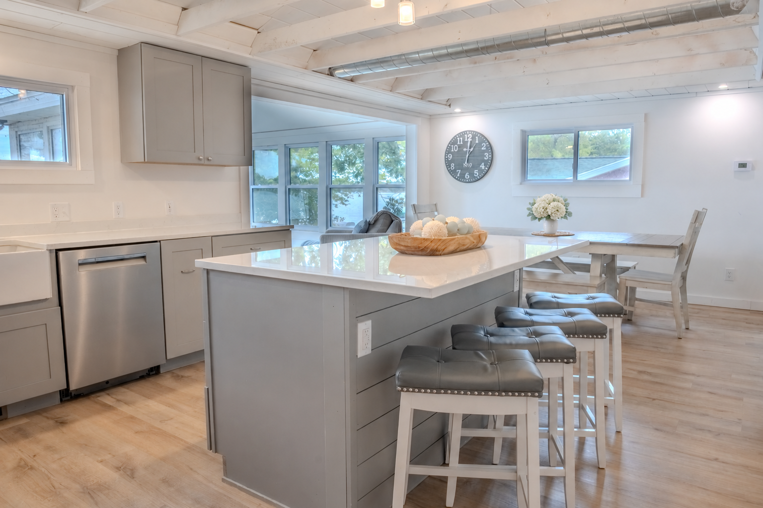 Bright modern kitchen with a large island, gray barstools, stainless steel dishwasher, dining table, and wall clock