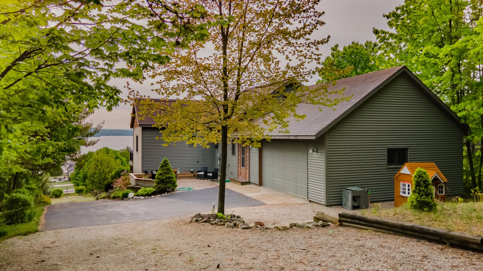Gray-sided suburban house with garage, driveway, landscaping, children’s playhouse, and wooded lake view