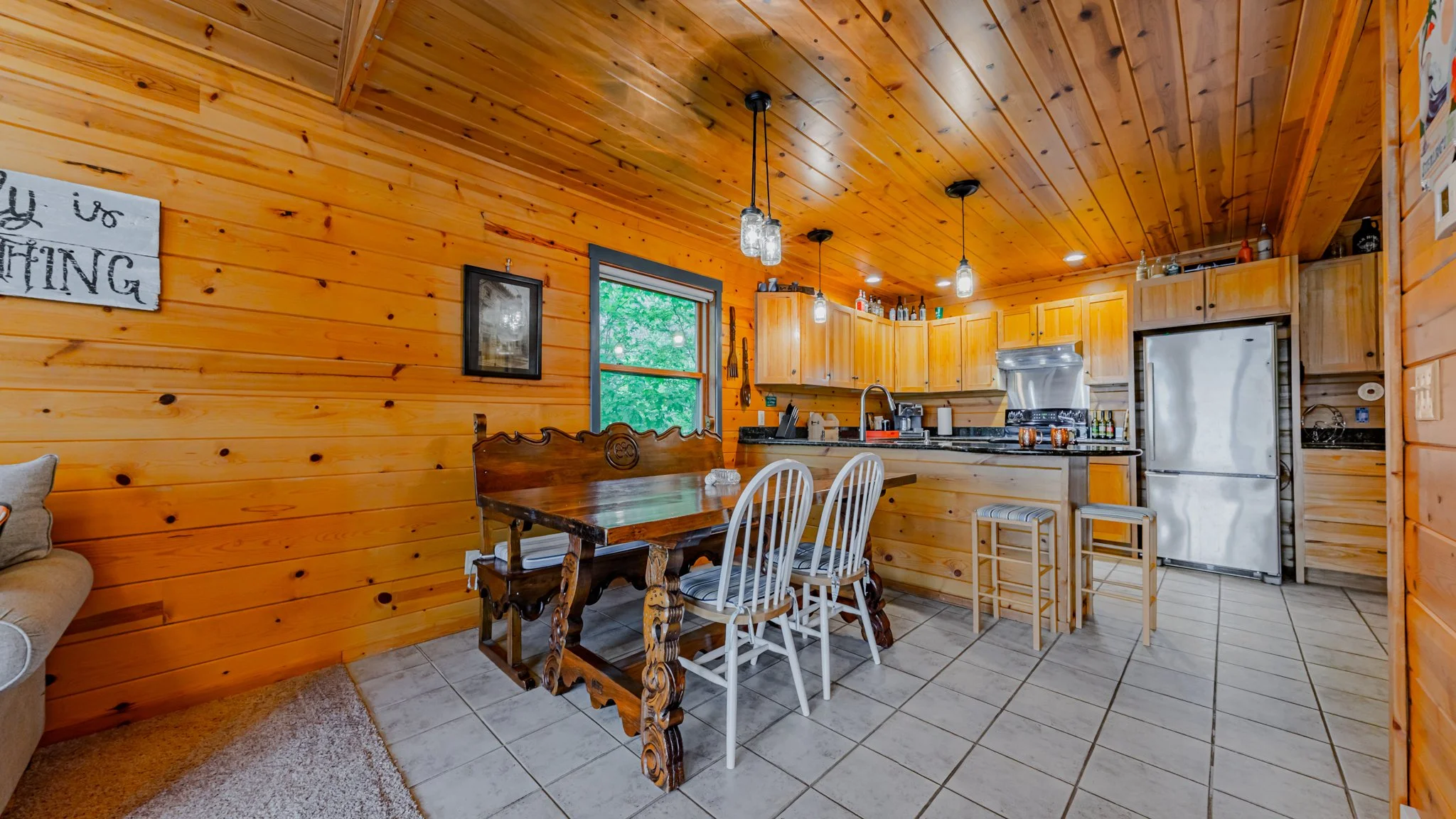 Rustic wood-paneled cabin kitchen with dining table, white chairs, wooden cabinets, stainless steel refrigerator, and tiled floor