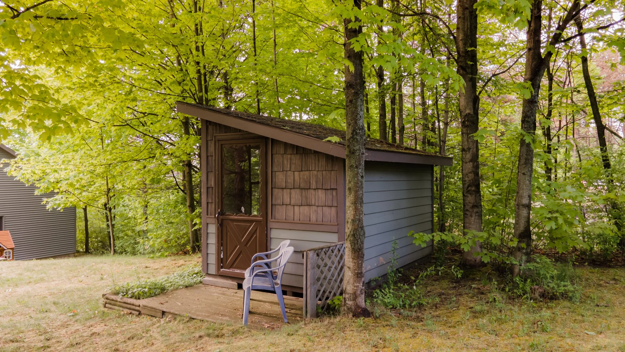 Small rustic cabin with a sloped roof, wooden door, deck, and plastic chairs in a leafy forest