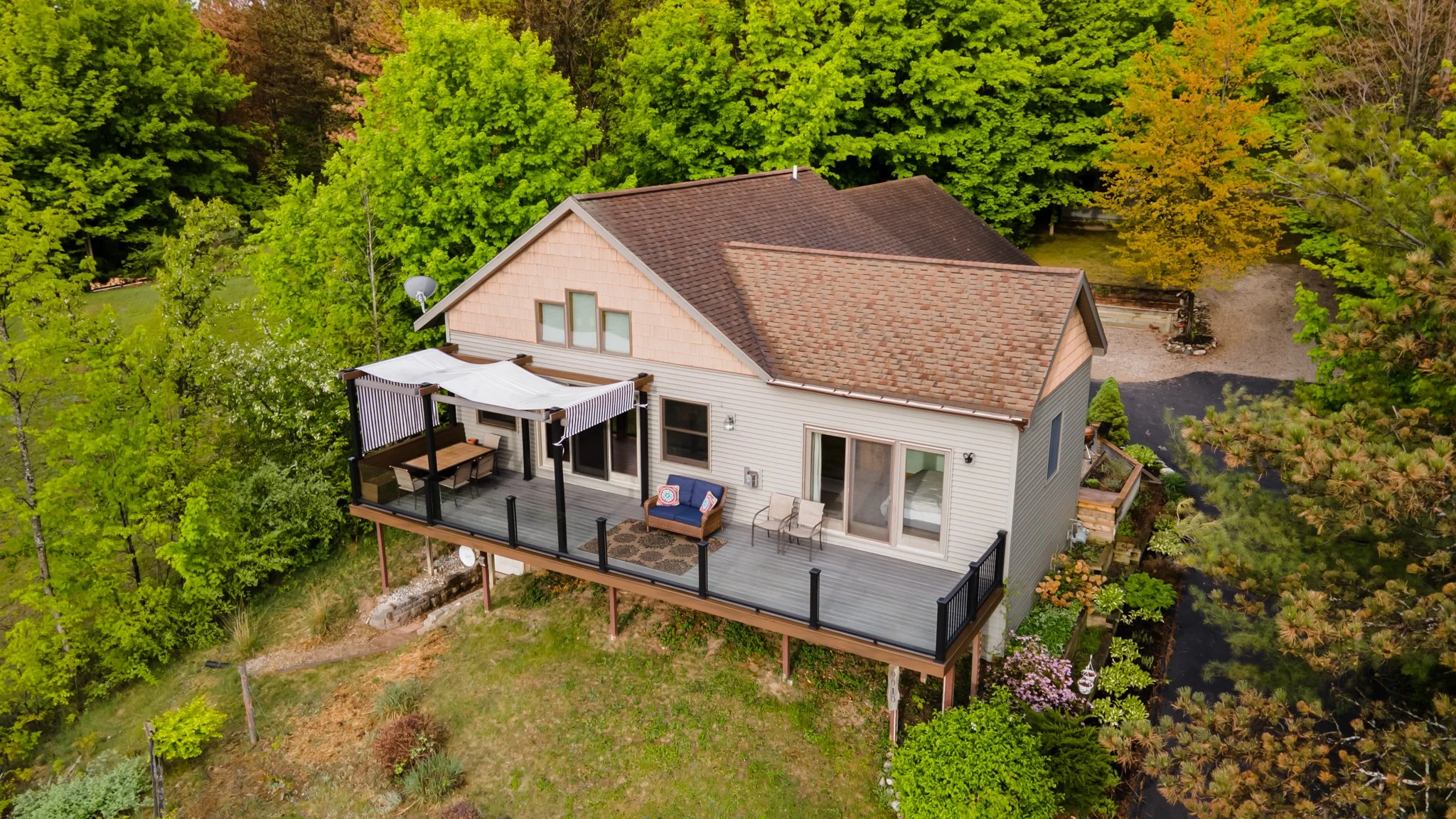 Aerial view of a suburban home with a large furnished deck surrounded by lush trees and landscaped gardens