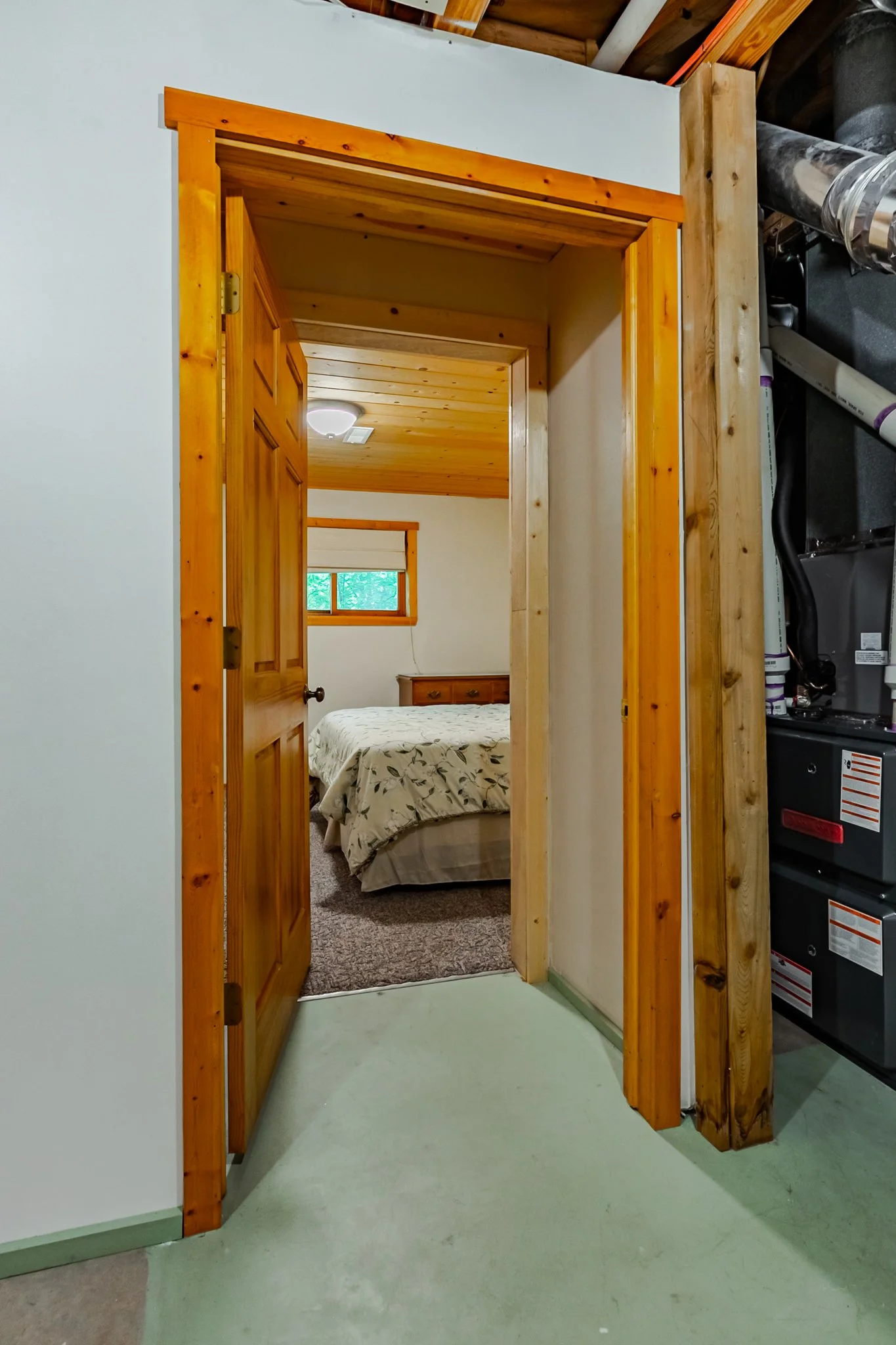 Open wooden doorway leading from a basement into a bedroom with a bed, window, and wood-paneled ceiling; furnace equipment is visible on the right.