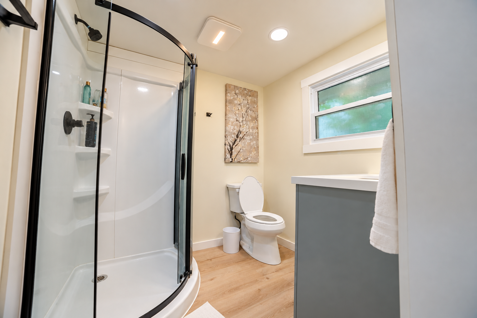 Bright modern bathroom with a curved glass shower, white toilet, gray vanity, window, and wood-look flooring