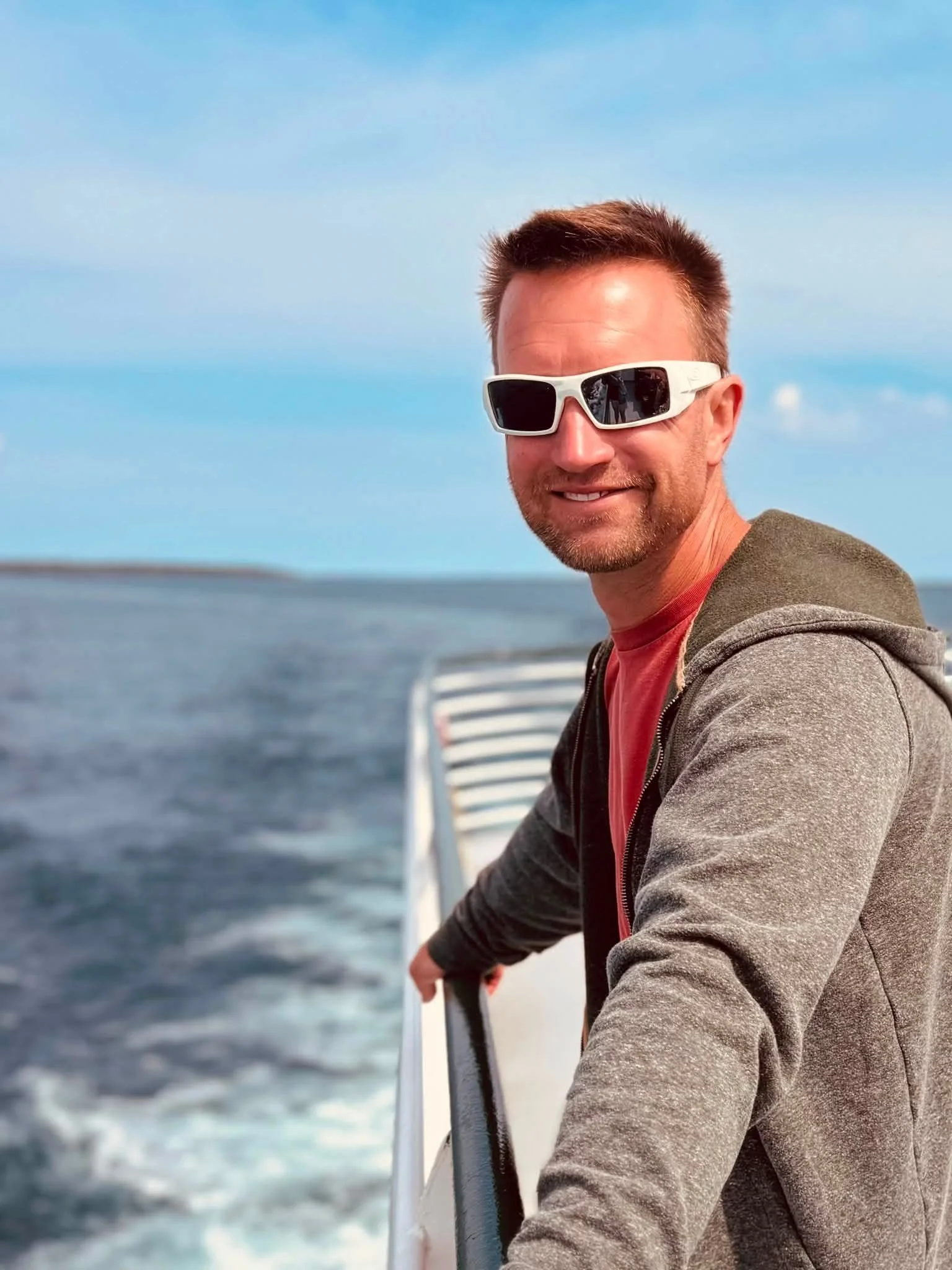 Man wearing white sunglasses and a gray hoodie standing on a boat over the ocean