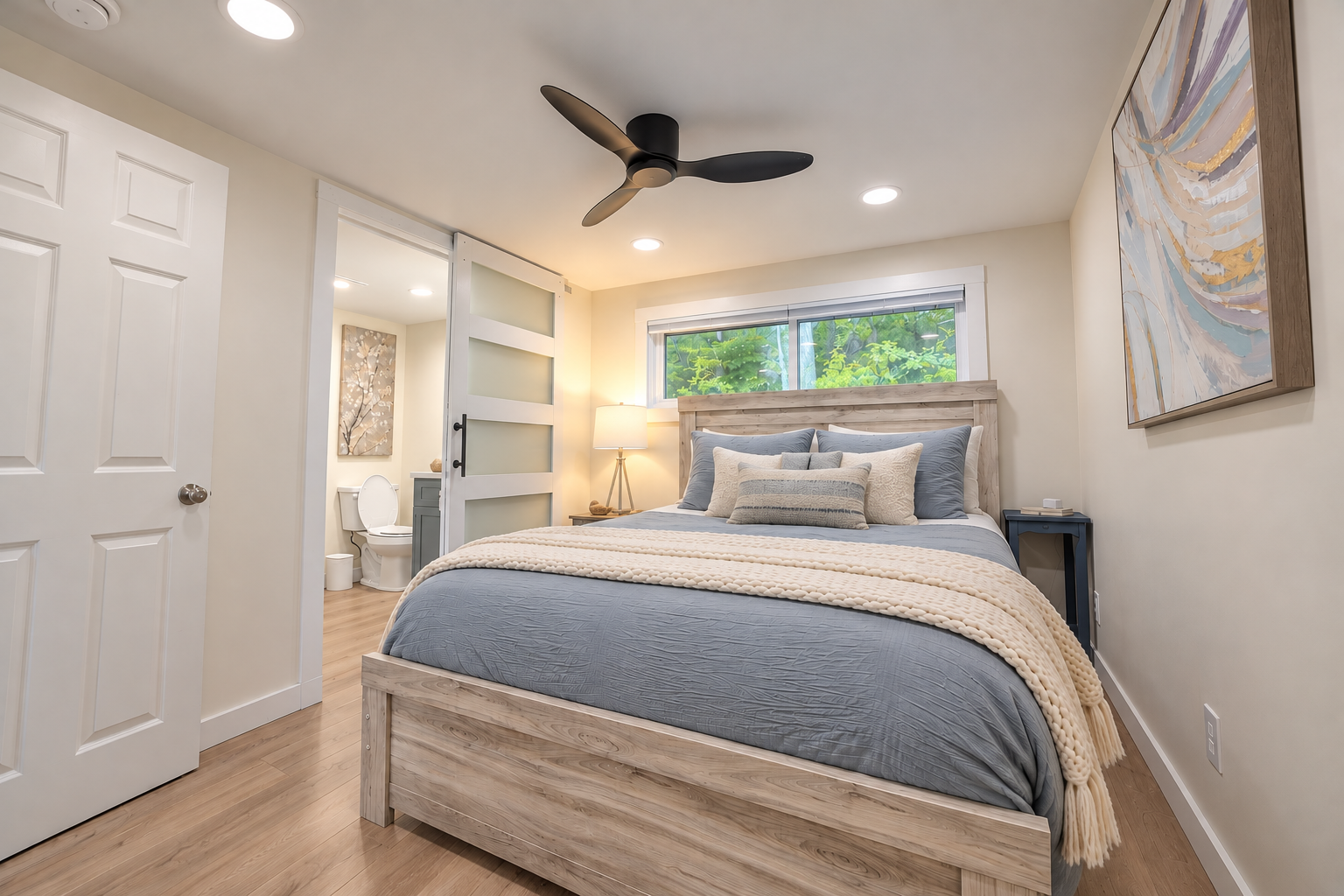 Bright modern bedroom with a wooden bed, blue bedding, cream throw blanket, ceiling fan, abstract wall art, and adjoining bathroom.