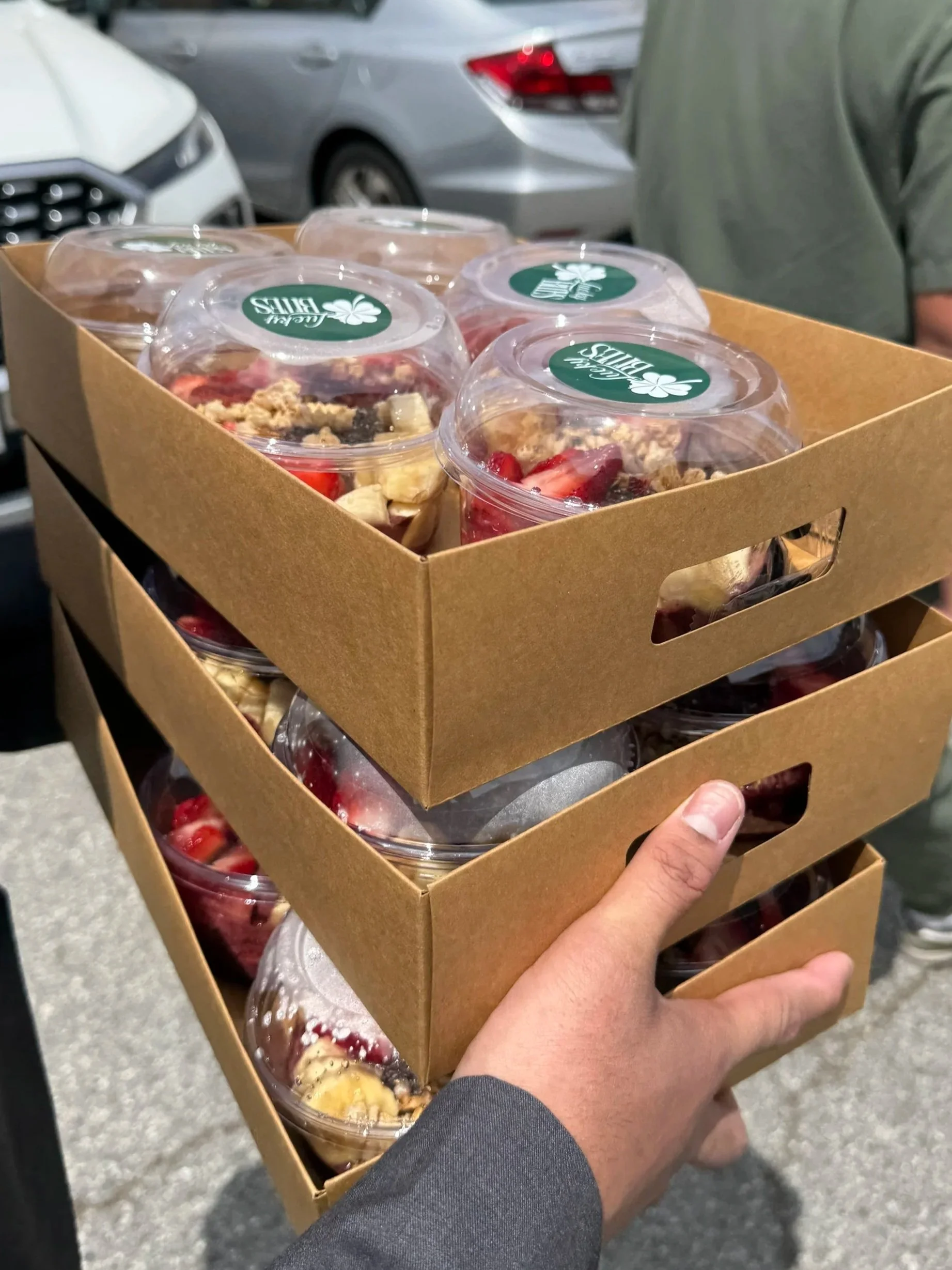 Hand holding a two-layer cardboard box with clear plastic cups of food, possibly parfaits or fruit salads, in a parking lot with cars in the background.