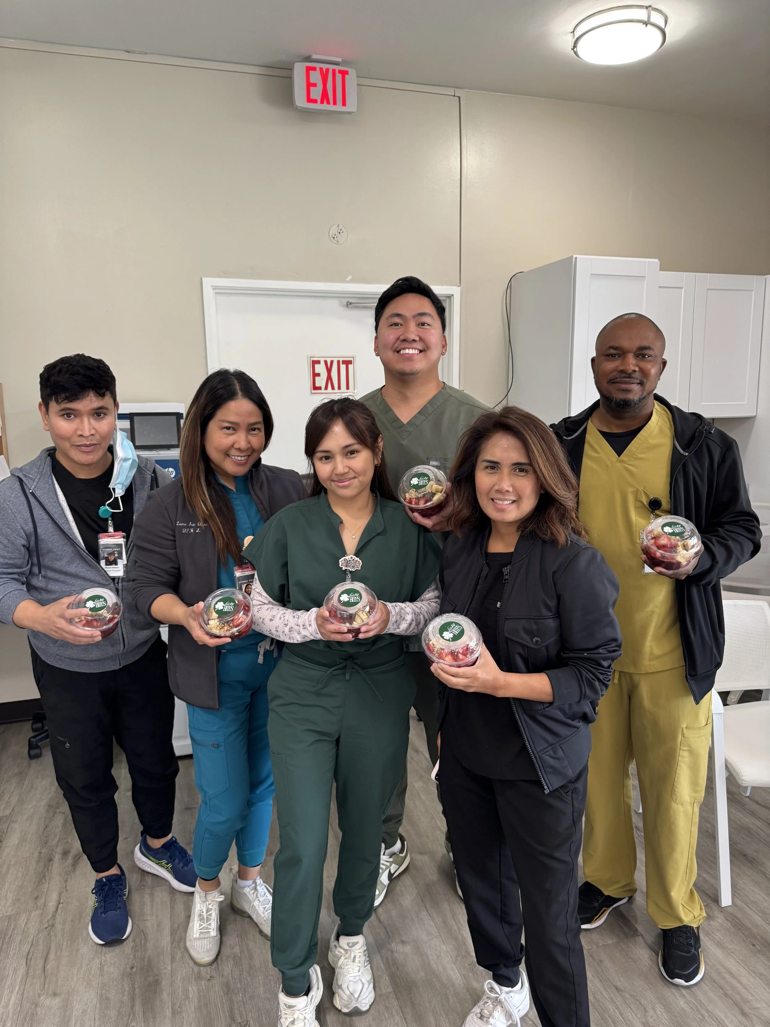 Group of six healthcare workers standing together indoors, holding bowls of fruit, smiling for the camera.